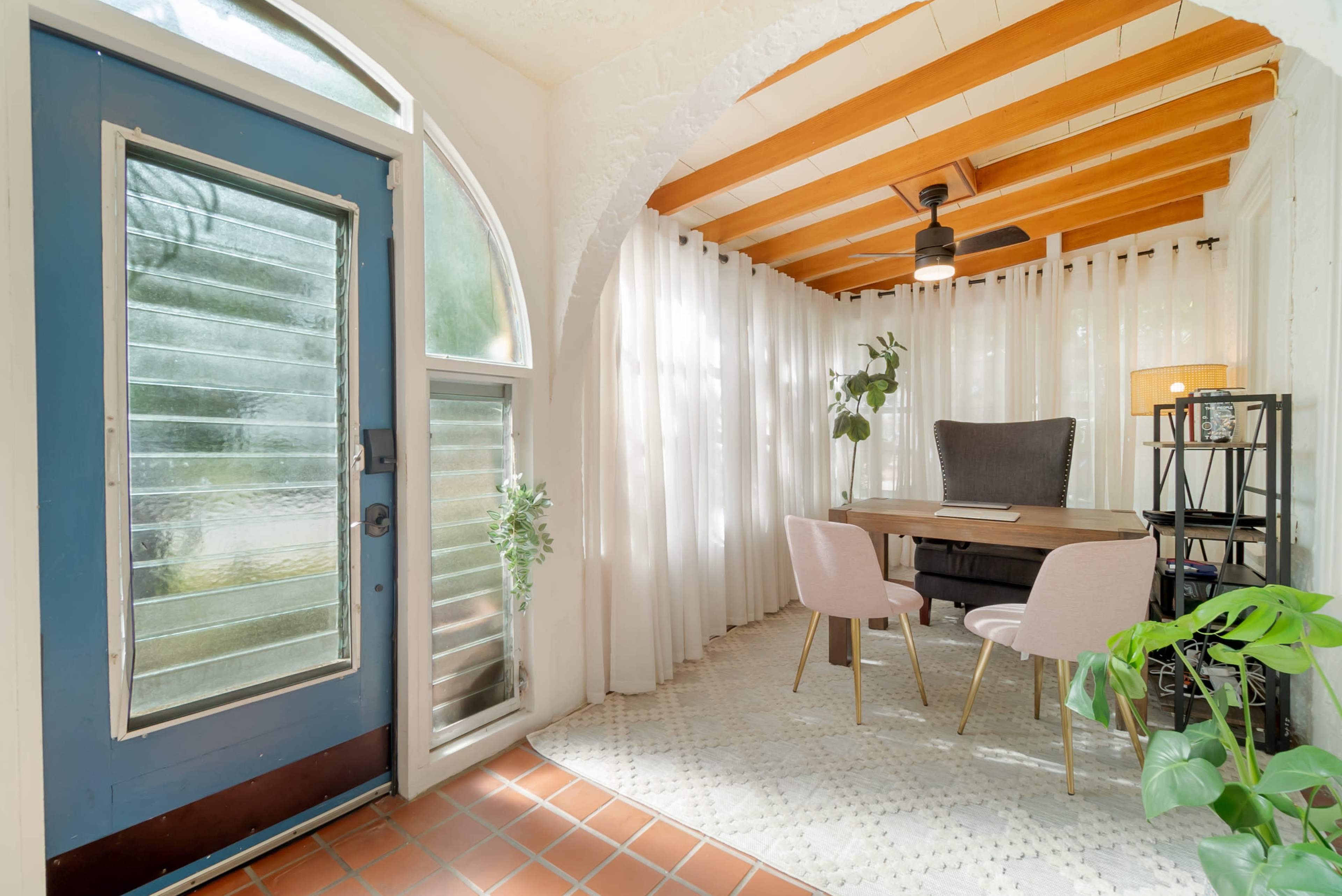 The image shows a well-lit interior space featuring a small dining table with pink chairs, large windows with white curtains, and a blue front door.