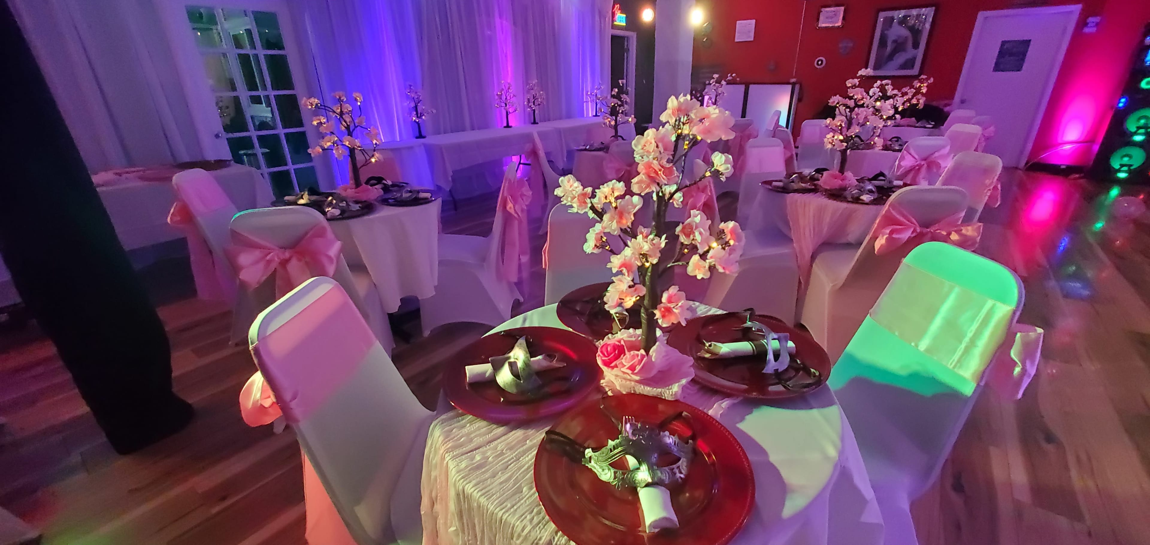 The scene depicts a decorated banquet hall with tables set for a formal event, featuring floral centerpieces and illuminated with colorful lighting.