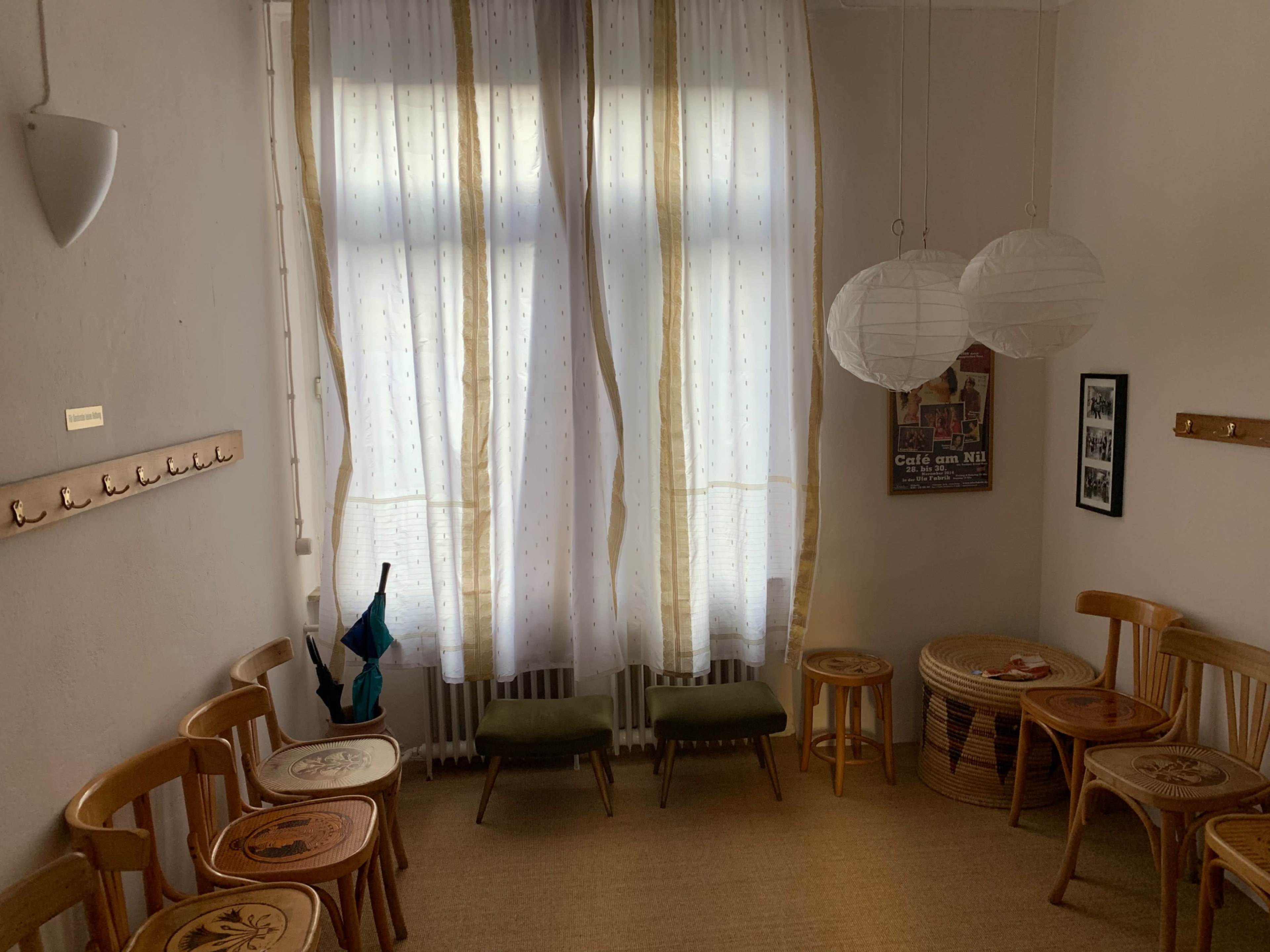 The room features several wooden chairs arranged around a neutral-colored floor, with two large windows covered by light curtains and hanging paper lanterns above.