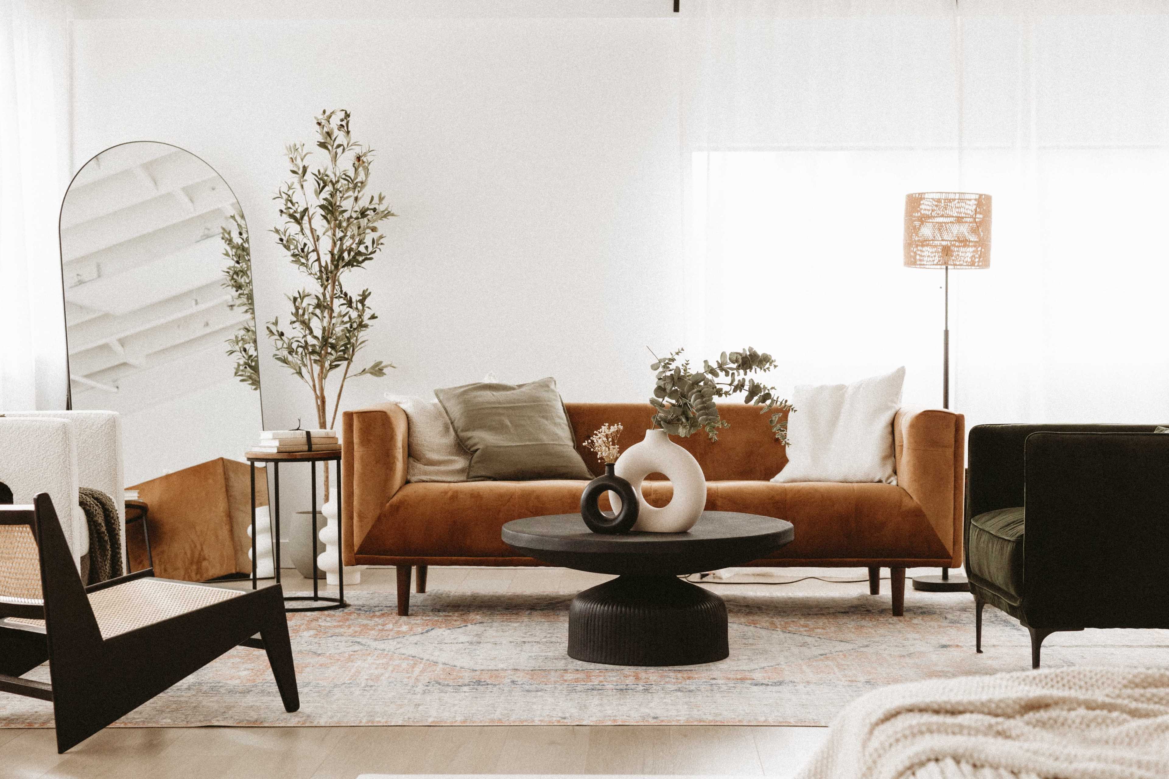 A modern living room with a brown velvet sofa, a round black coffee table, a green armchair, and decorative plants.
