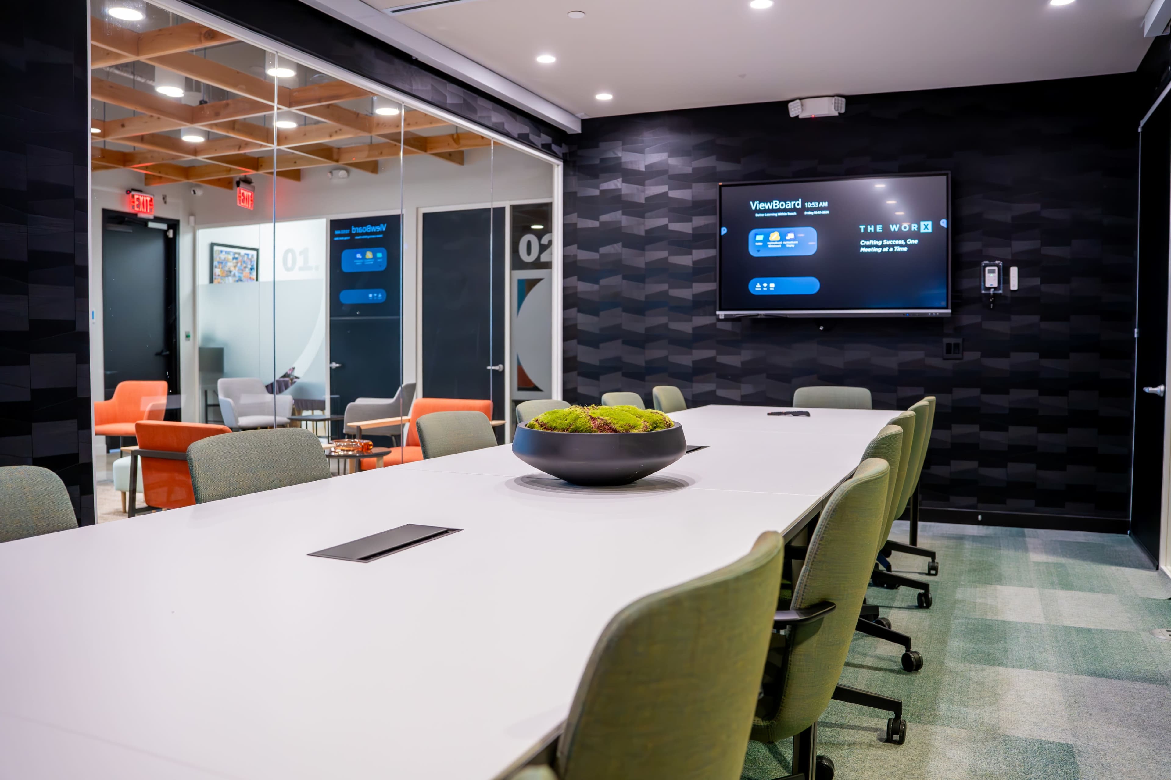 A modern conference room features a long white table, comfortable green chairs, and a large screen displaying the VineBoard interface, with a bowl of greenery at the center.