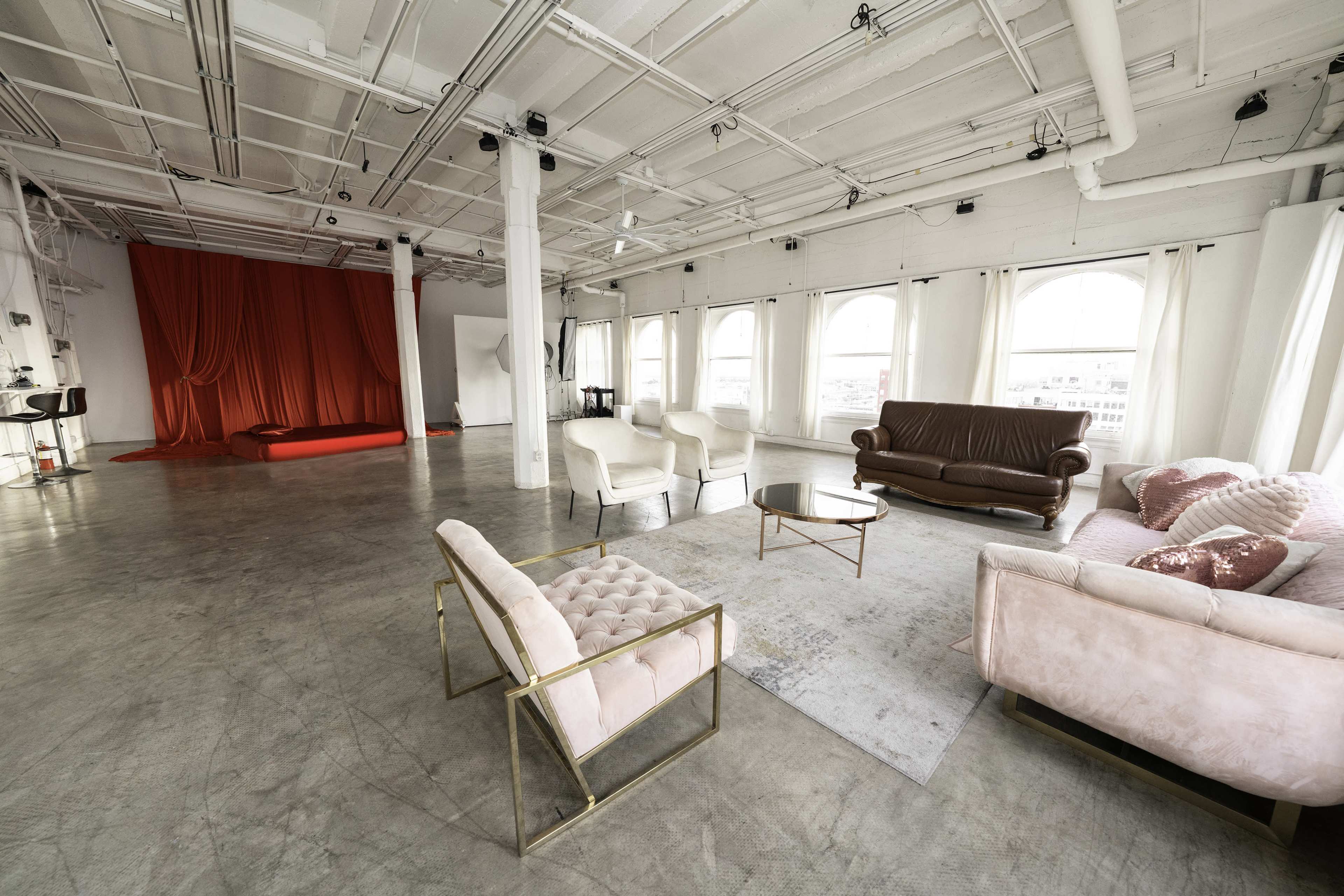 The image shows a spacious, brightly lit studio with a seating area featuring various sofas and chairs, along with a red backdrop at one end.
