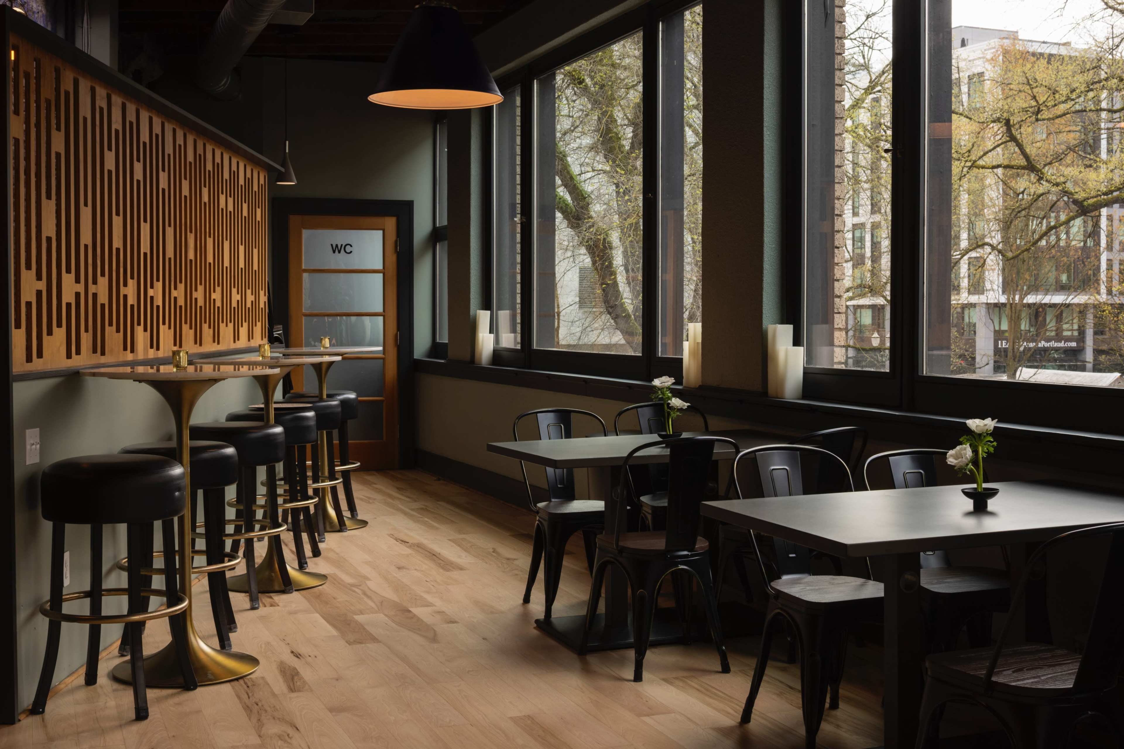 The image shows a modern café interior with wooden flooring, black metal furniture, and large windows allowing natural light to fill the space.