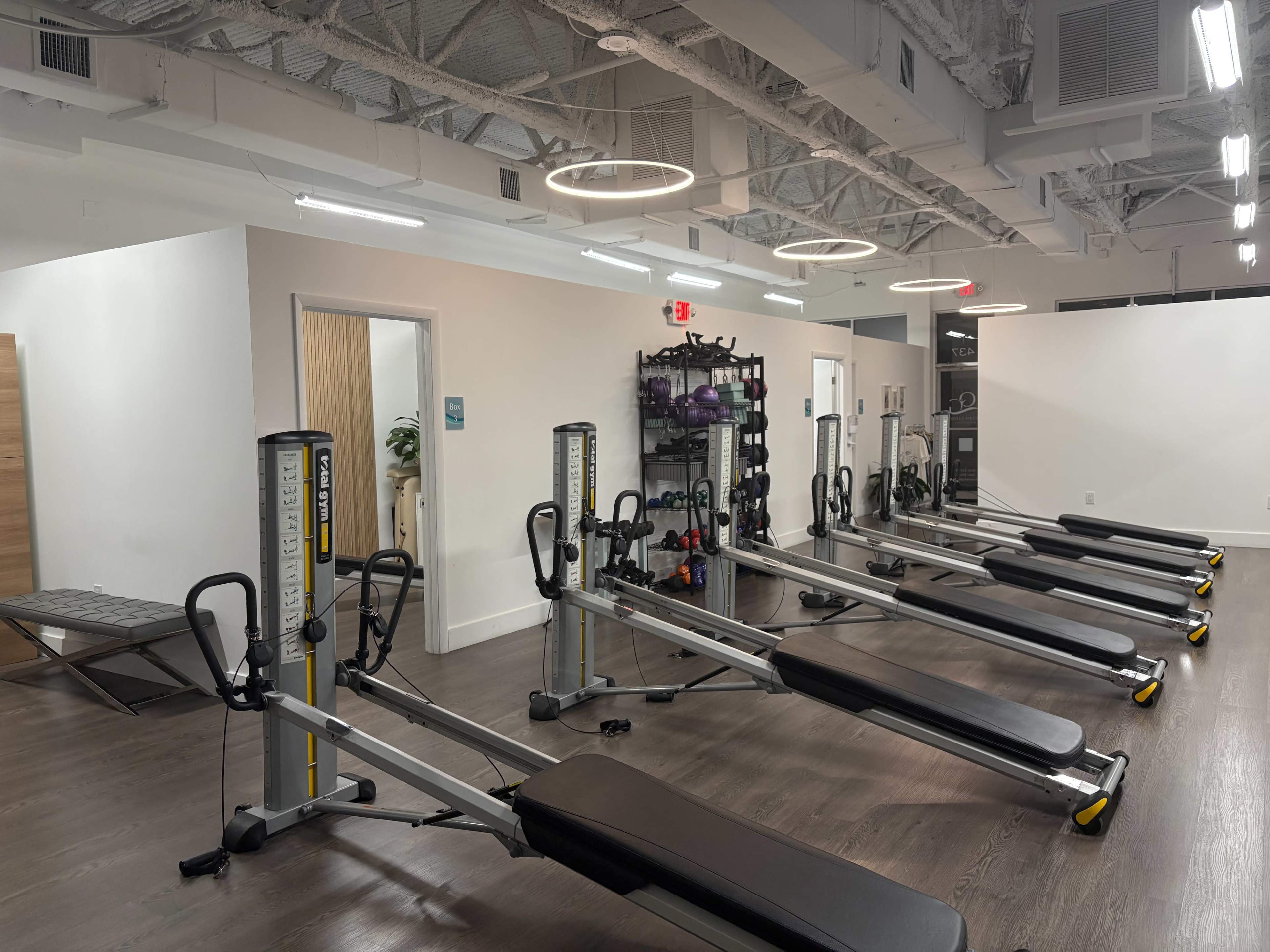 The image shows a fitness studio with several reformer machines arranged in a row on a wooden floor, alongside shelves filled with exercise equipment.