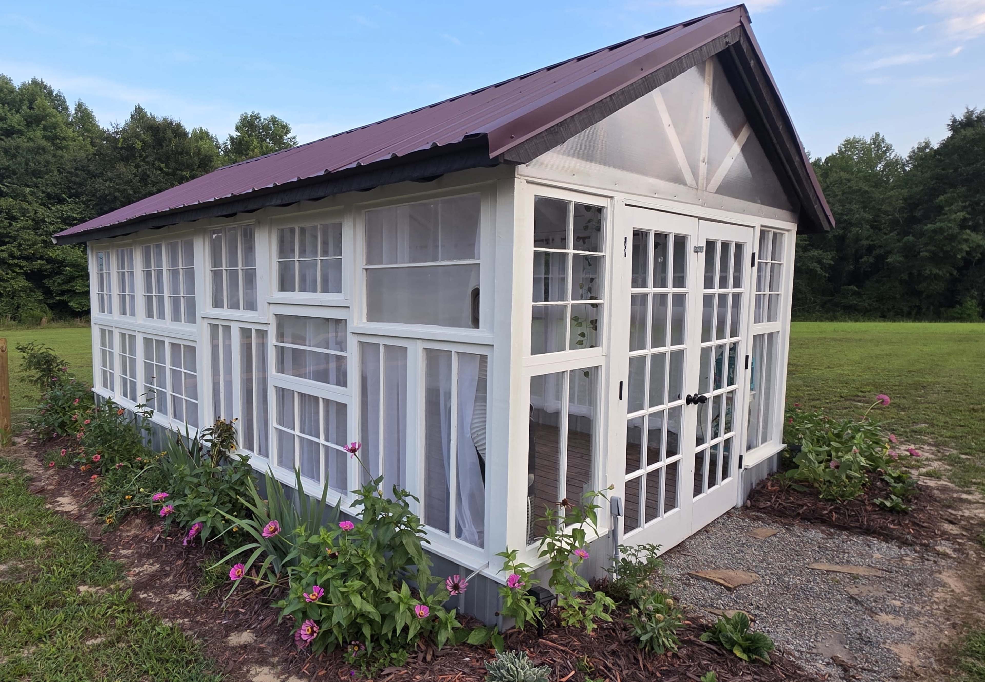 The image shows a small greenhouse with multiple windows and double doors, surrounded by flowering plants and located in a grassy area.