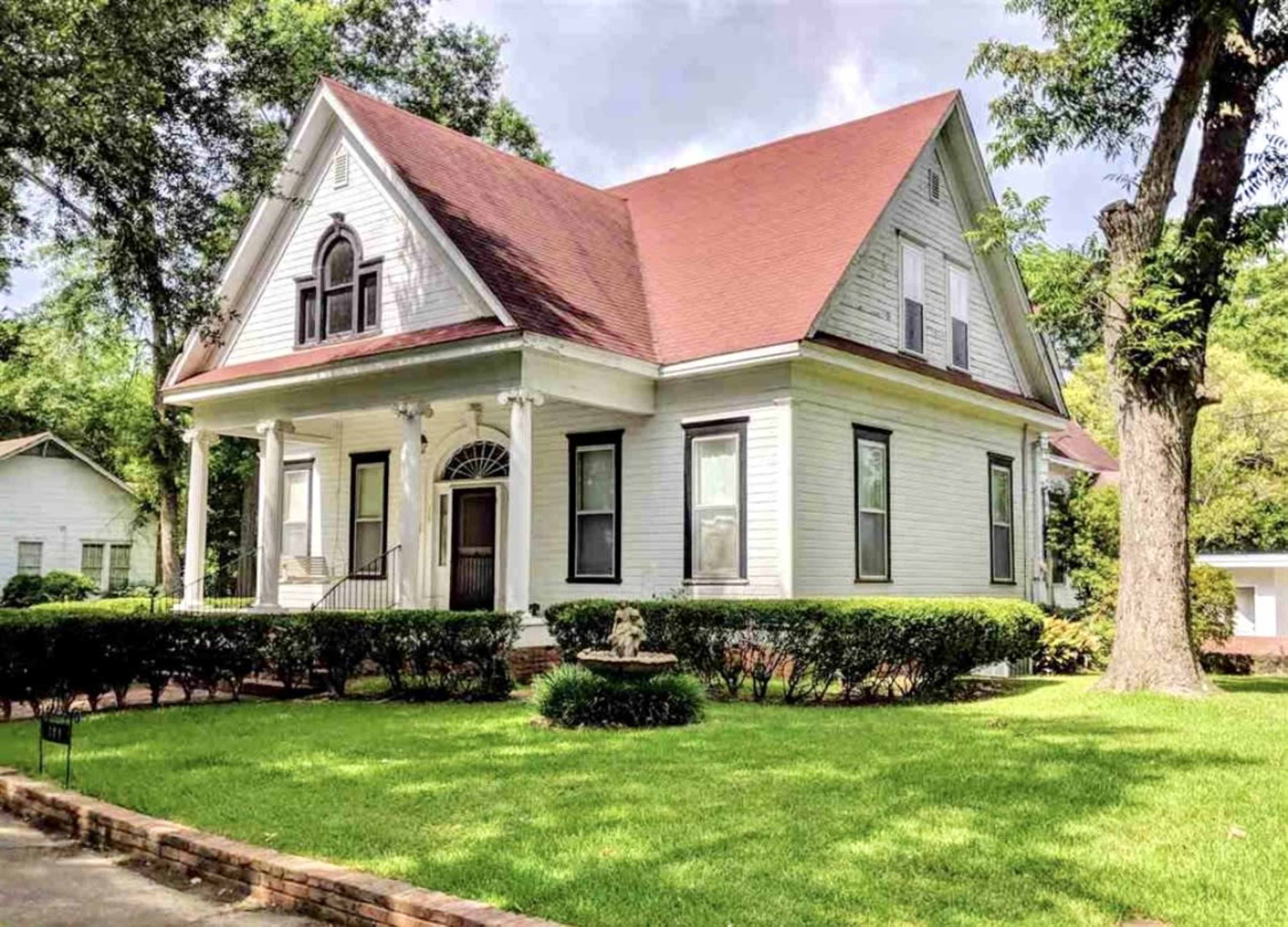 A large white house with a red roof features a front porch and manicured green lawns, surrounded by low hedges and trees.