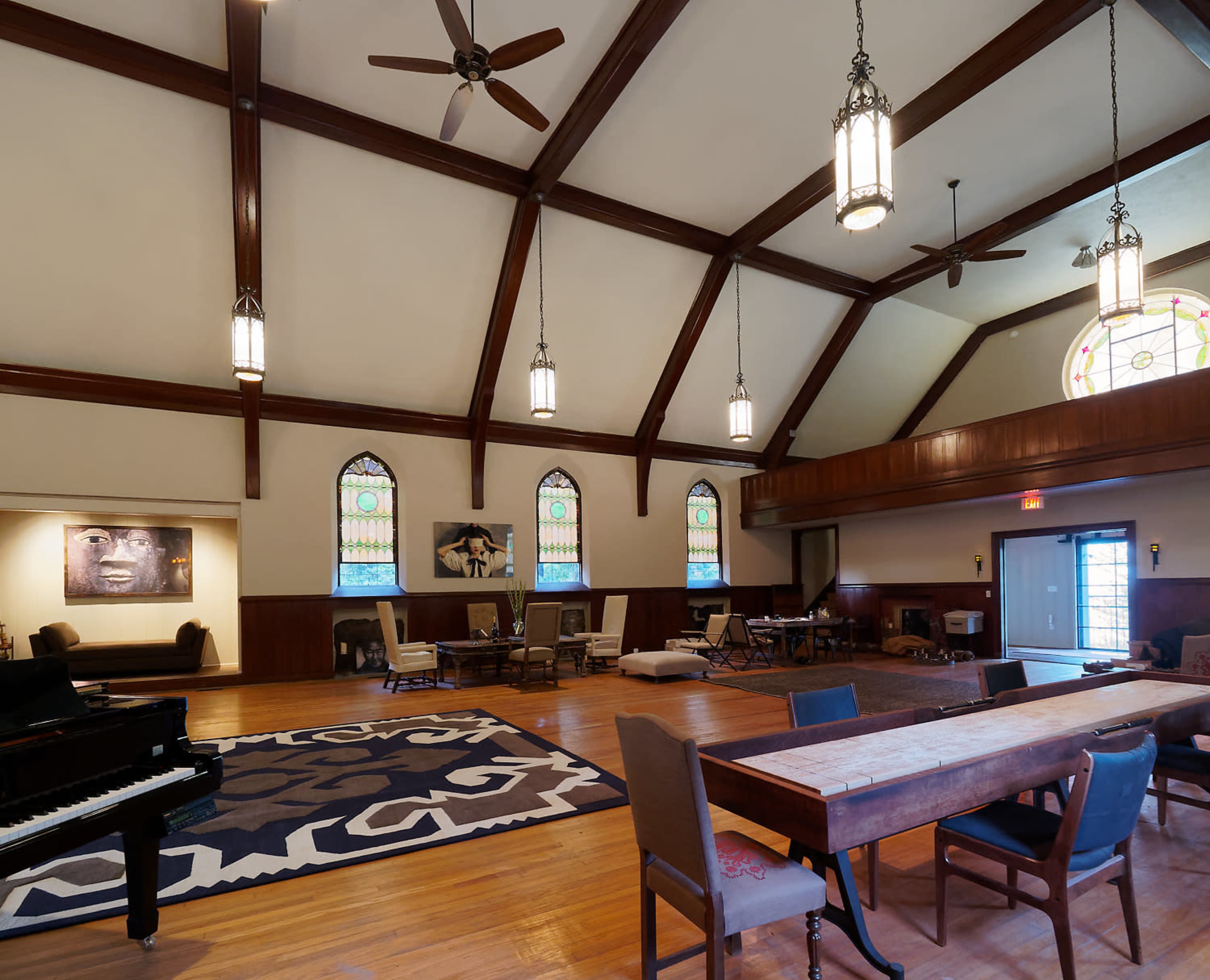 A spacious room with high ceilings, wooden beams, large stained glass windows, and a mix of seating arrangements, including a piano and a long dining table.