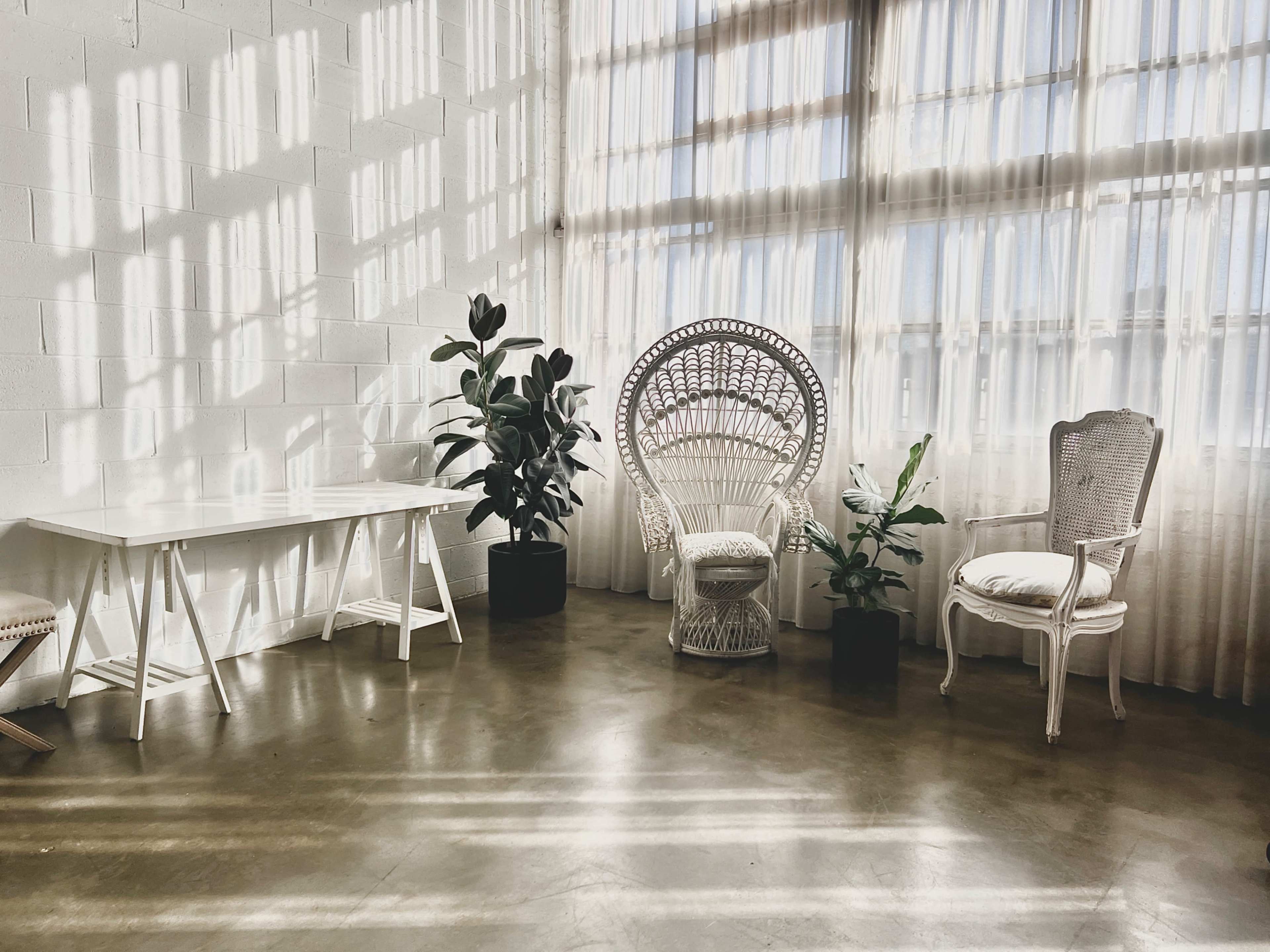 A well-lit room features a large window with sheer curtains, a white table, a decorative chair, and two potted plants.