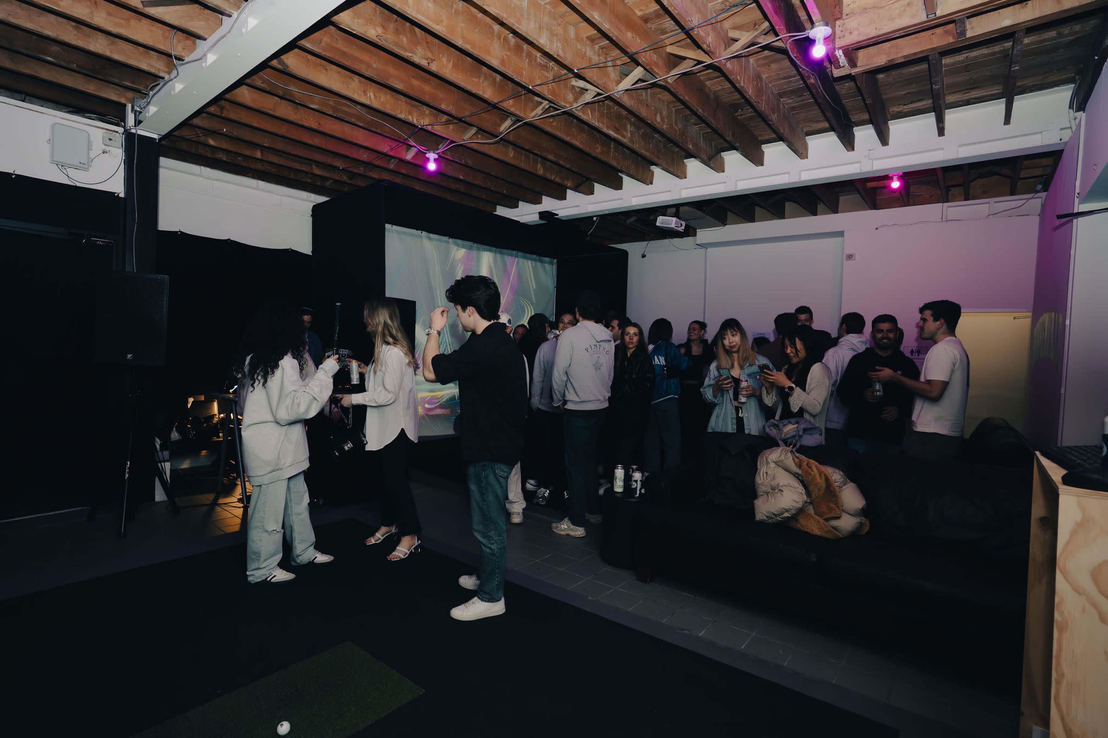 A group of people socialize and dance in a dimly lit room with colorful projections on the wall and a sofa in the background.