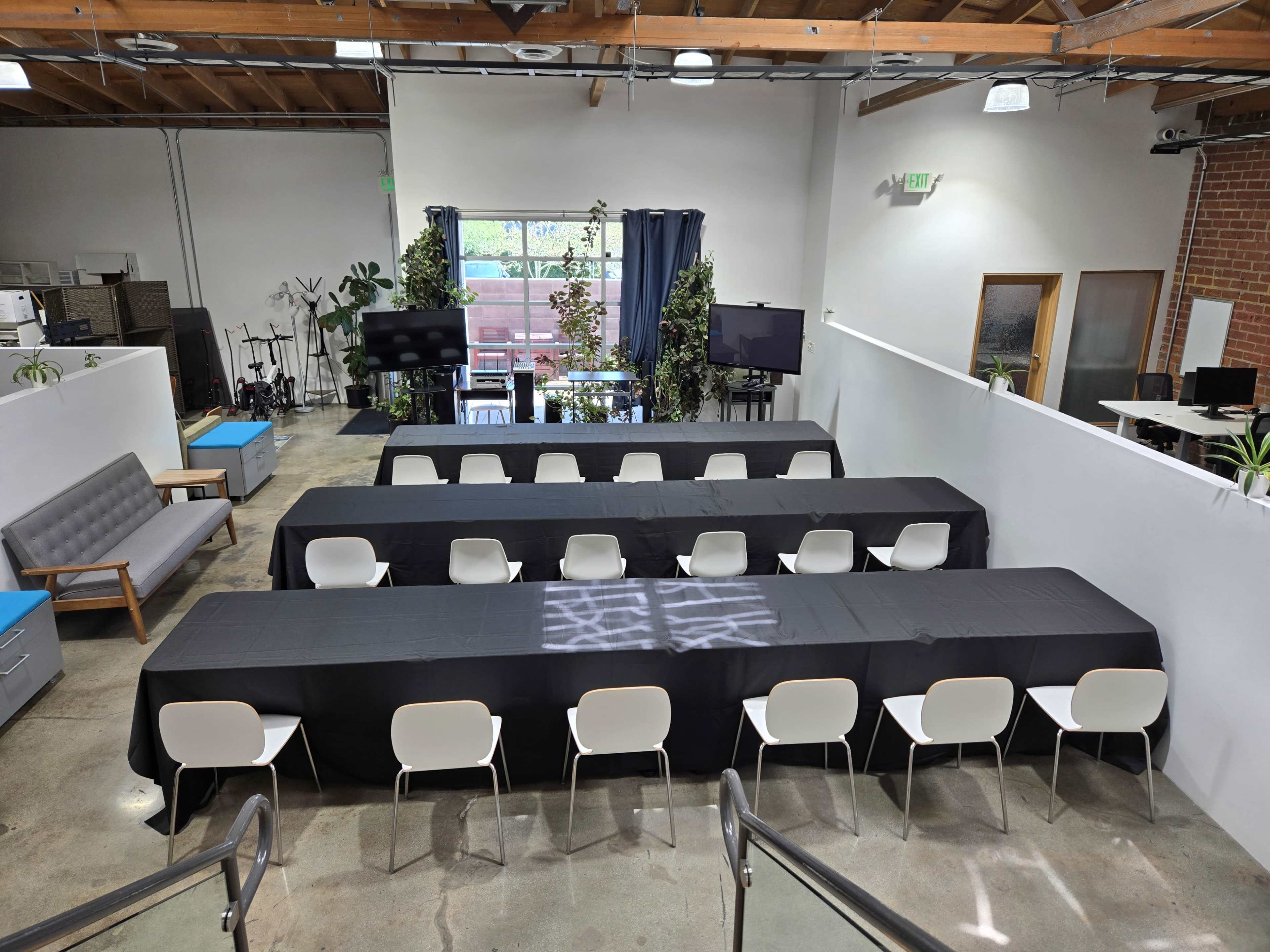 The image shows a modern workspace featuring a large arrangement of tables covered in black tablecloths, surrounded by white chairs, with large windows and greenery in the background.