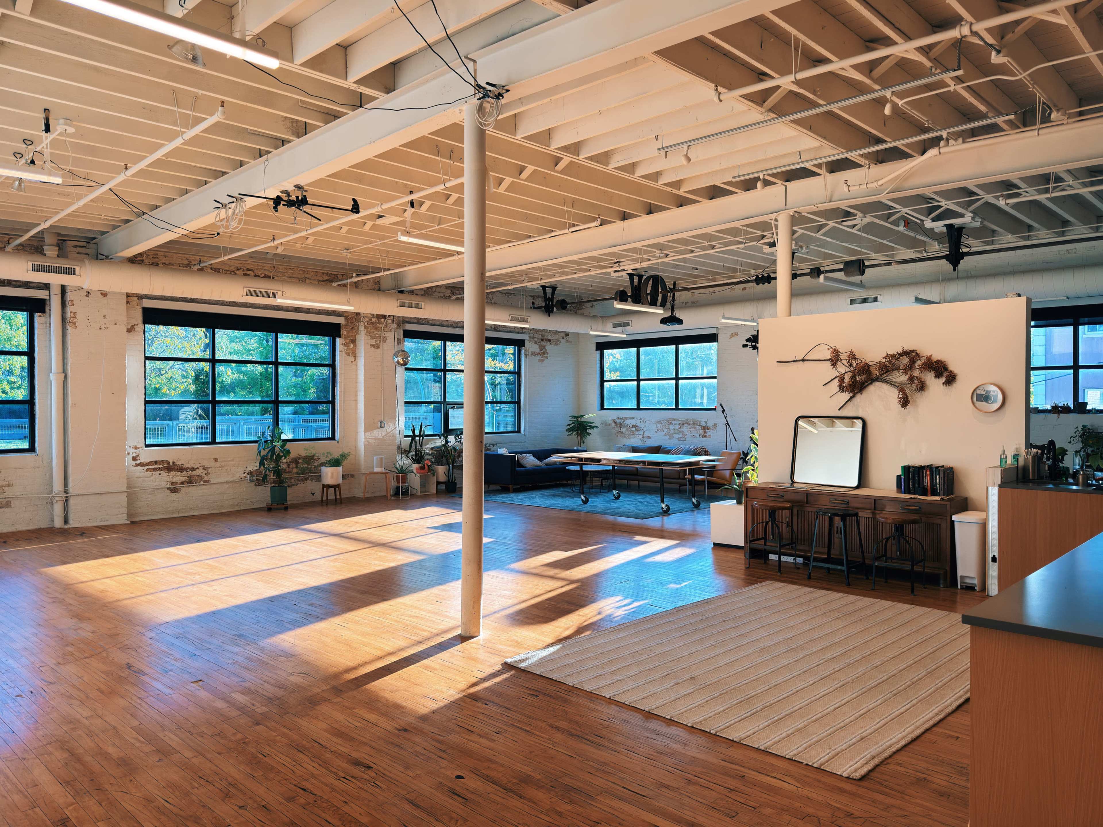 The image features a spacious, well-lit interior of a modern loft with large windows, wooden floors, and minimalist furnishings.