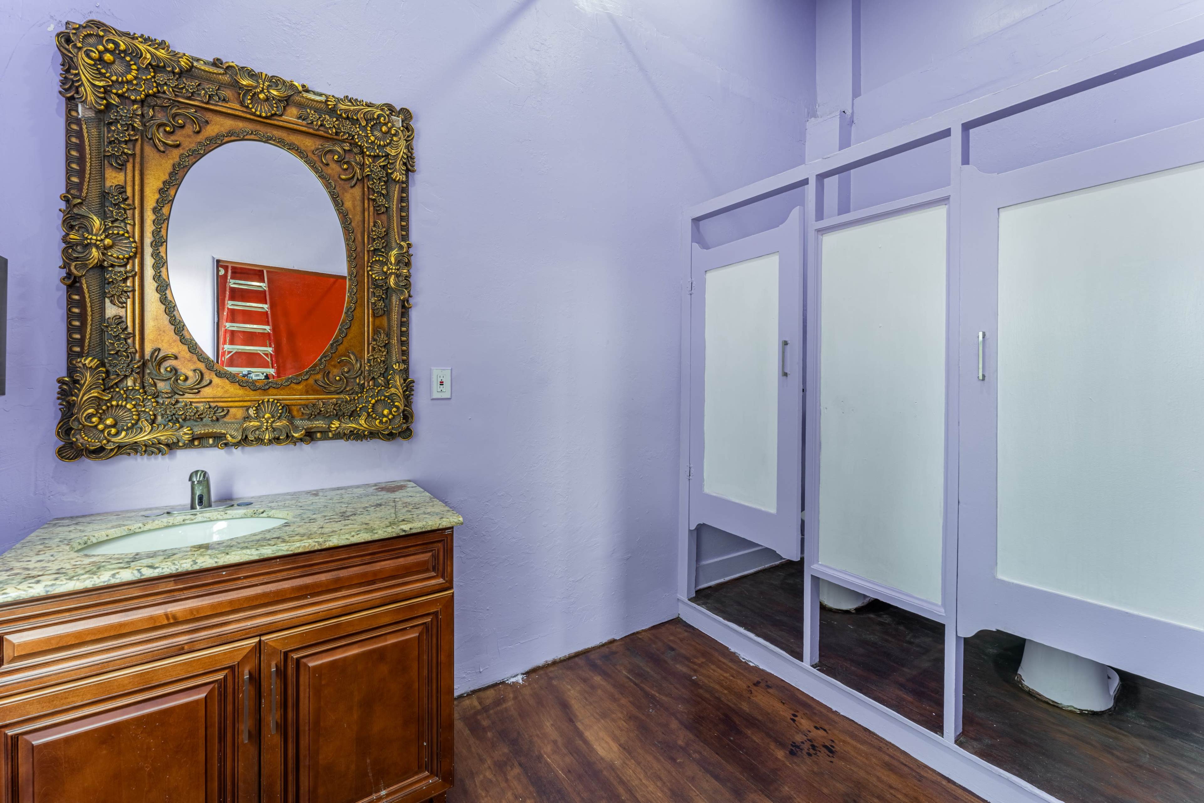 A bathroom with a wooden vanity and sink, a large ornate mirror, and three frosted glass shower doors against a lavender wall.
