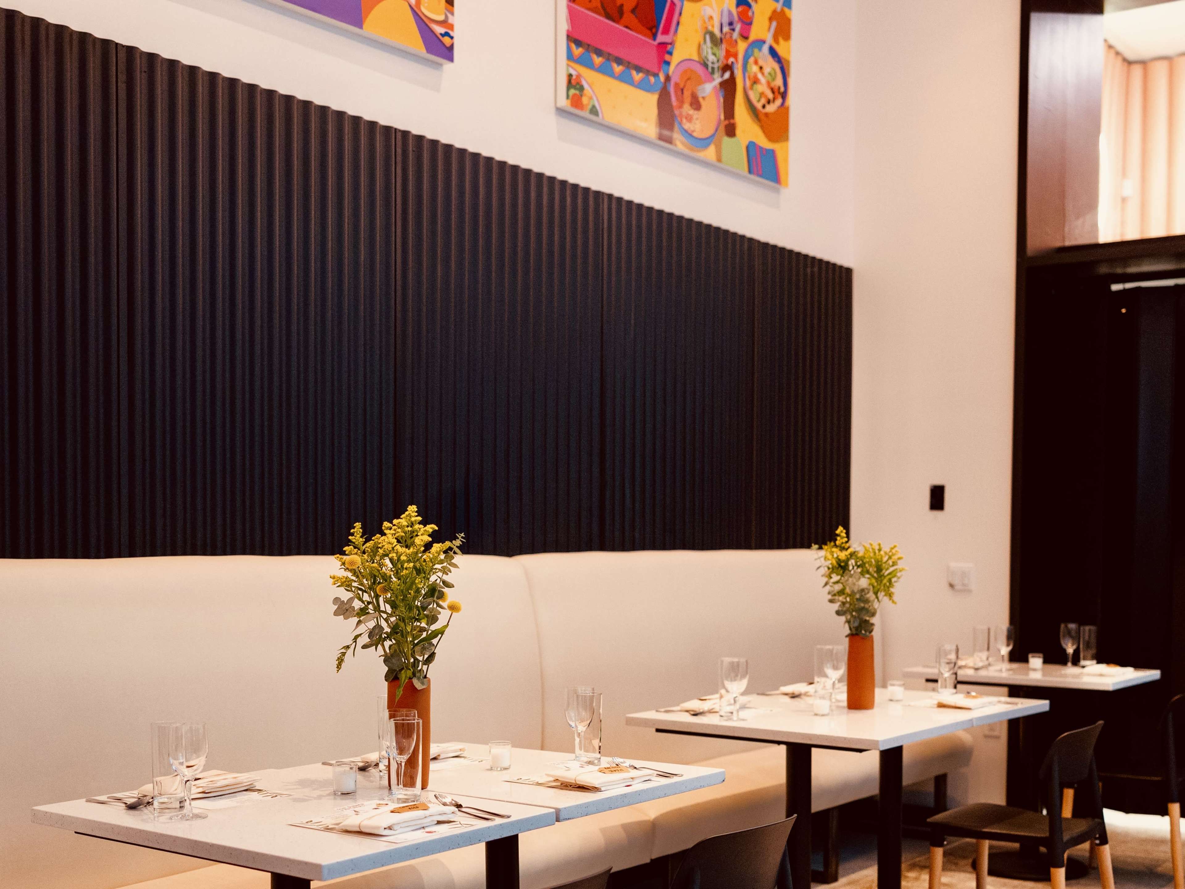 A dining area in a restaurant with neatly set tables, each featuring a small vase of flowers, against a backdrop of textured black walls and colorful artwork.