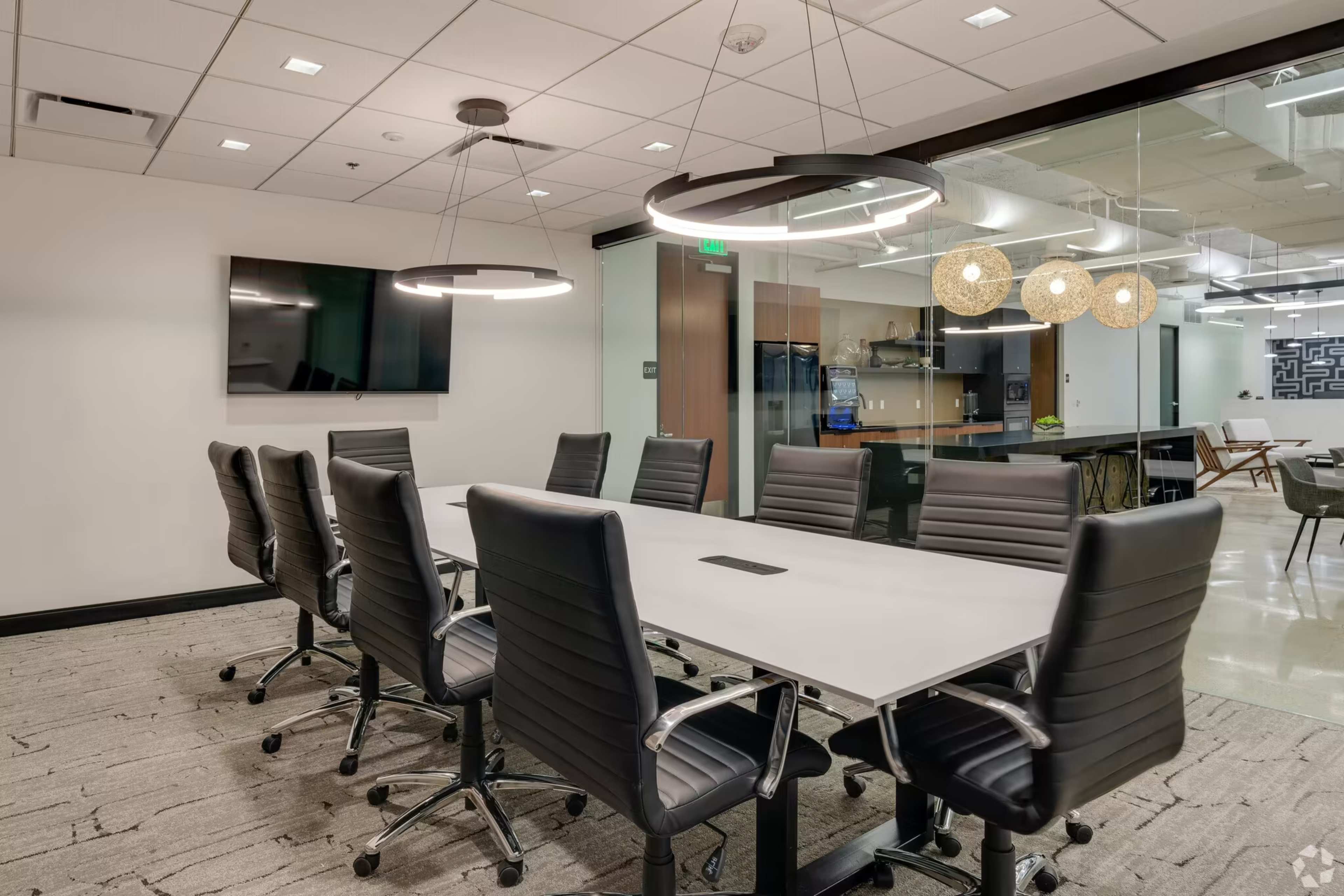 The image shows a modern conference room with a long table, black chairs, and a large screen, adjacent to an open kitchen area.