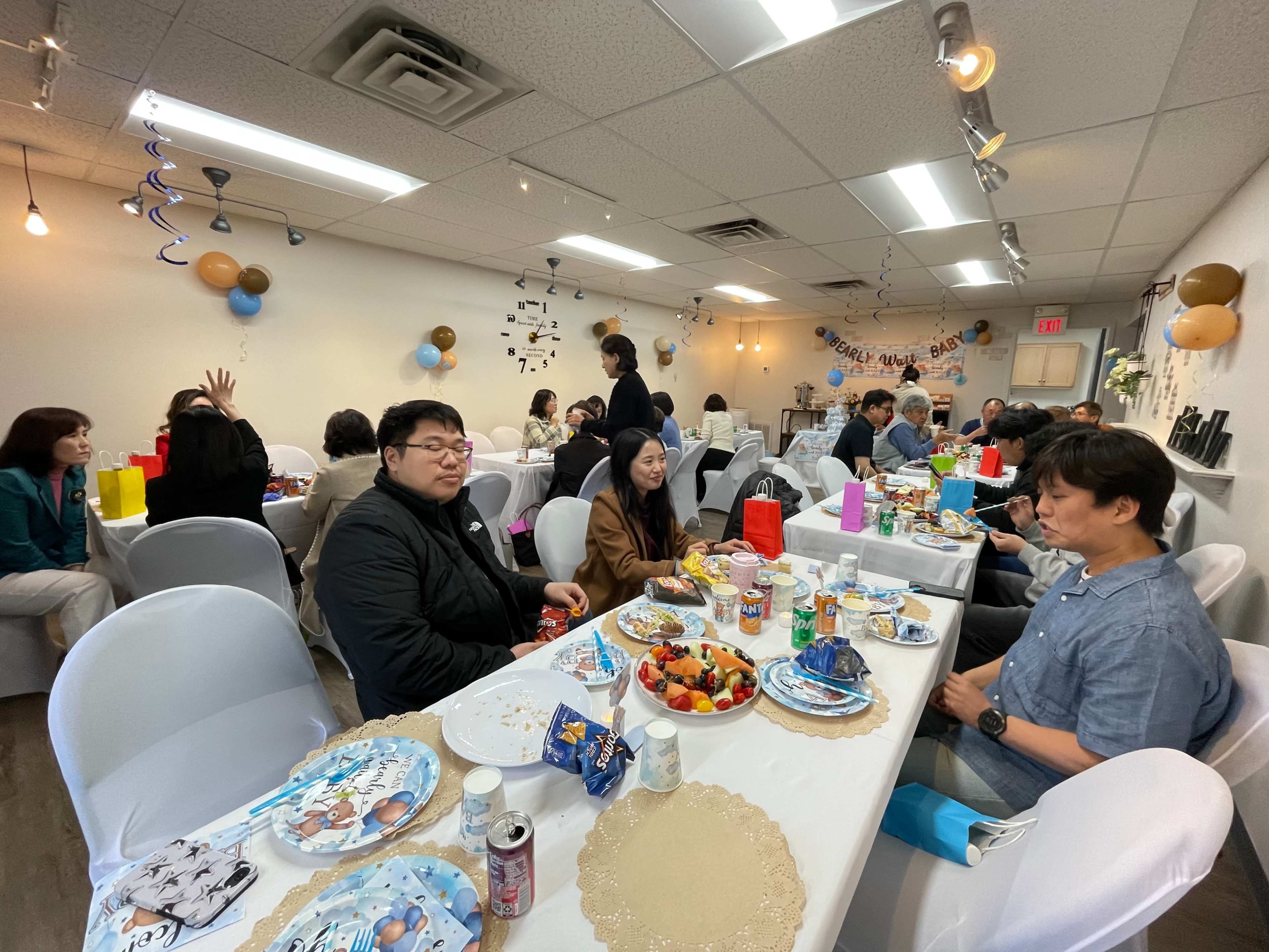 A group of people is seated at tables in a decorated room, enjoying food and drinks during an event or gathering.