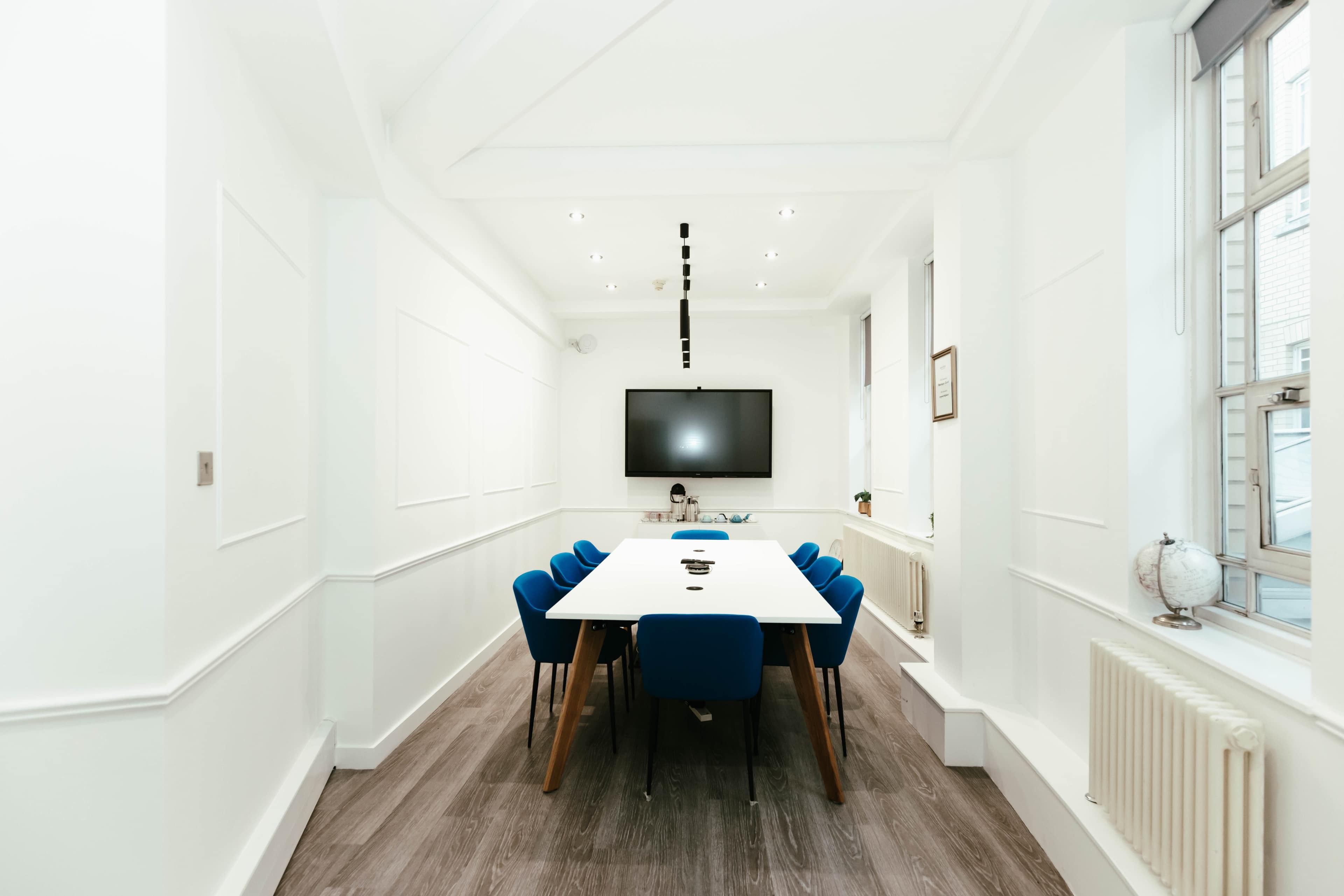 A modern conference room features a long white table surrounded by blue chairs, with a large flat-screen TV mounted on the wall.