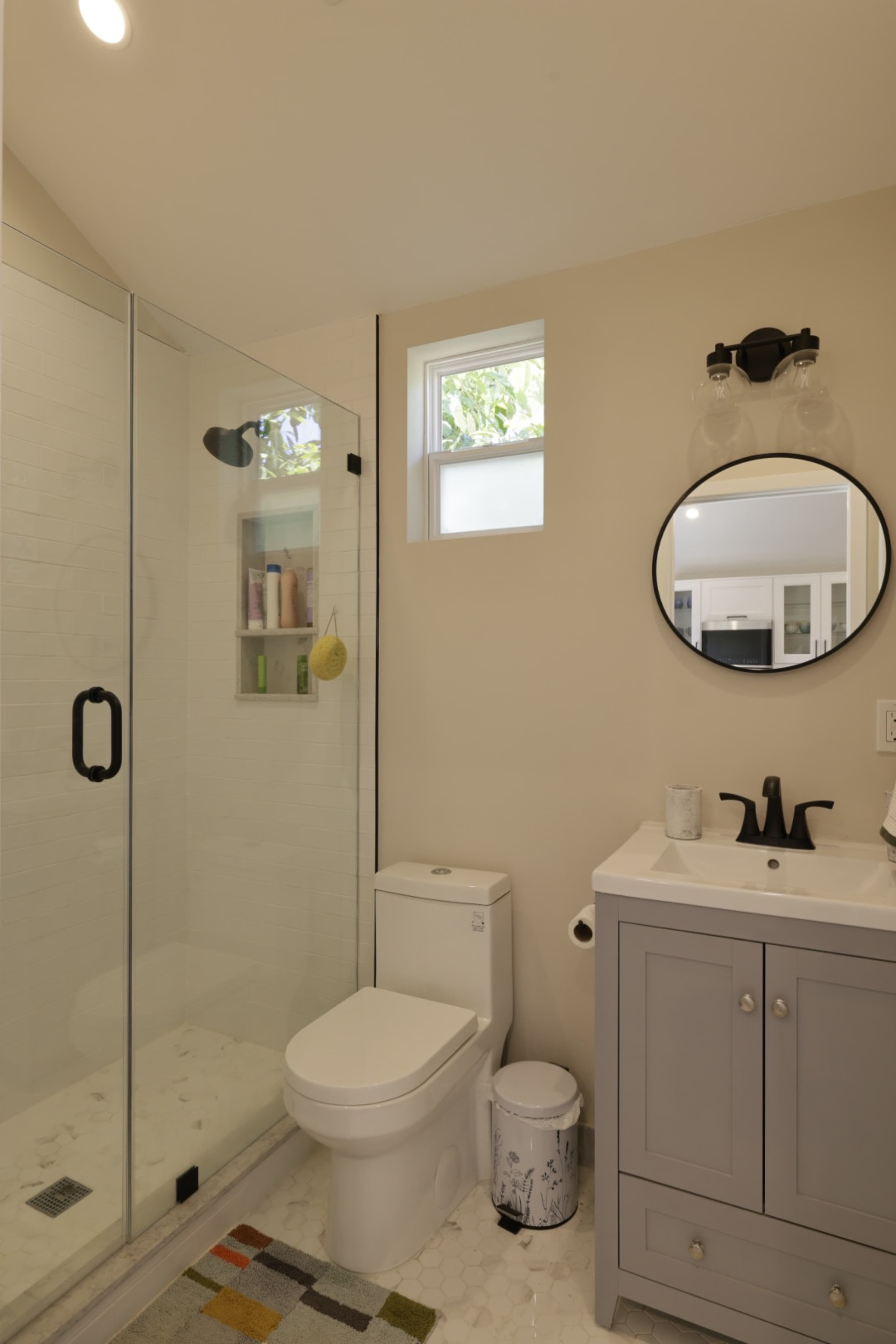 A modern bathroom with a glass shower enclosure, a round mirror above a grey vanity, and a white toilet.