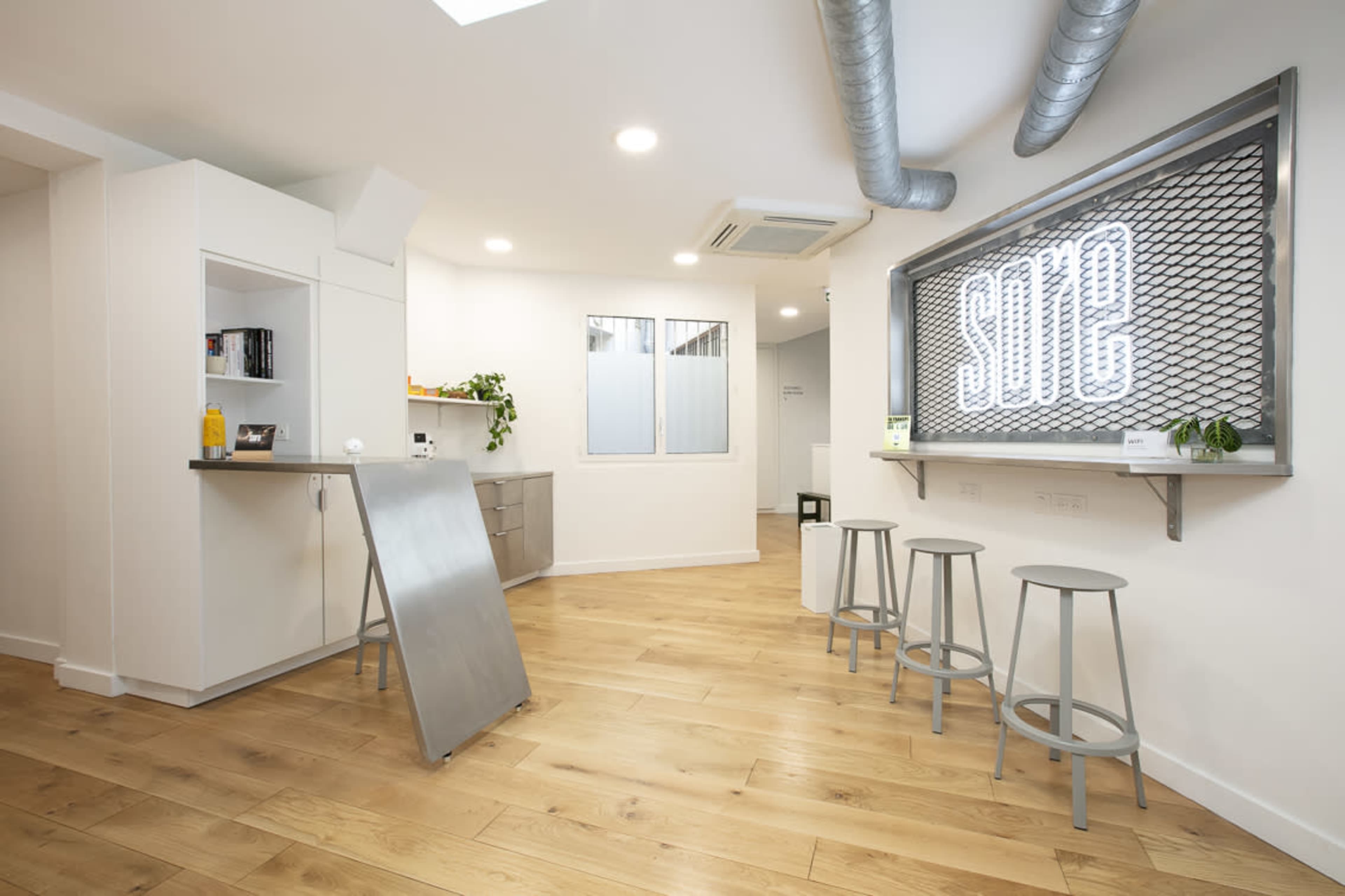 A modern interior space with light wood flooring, white walls, and a bar area with three metal stools.
