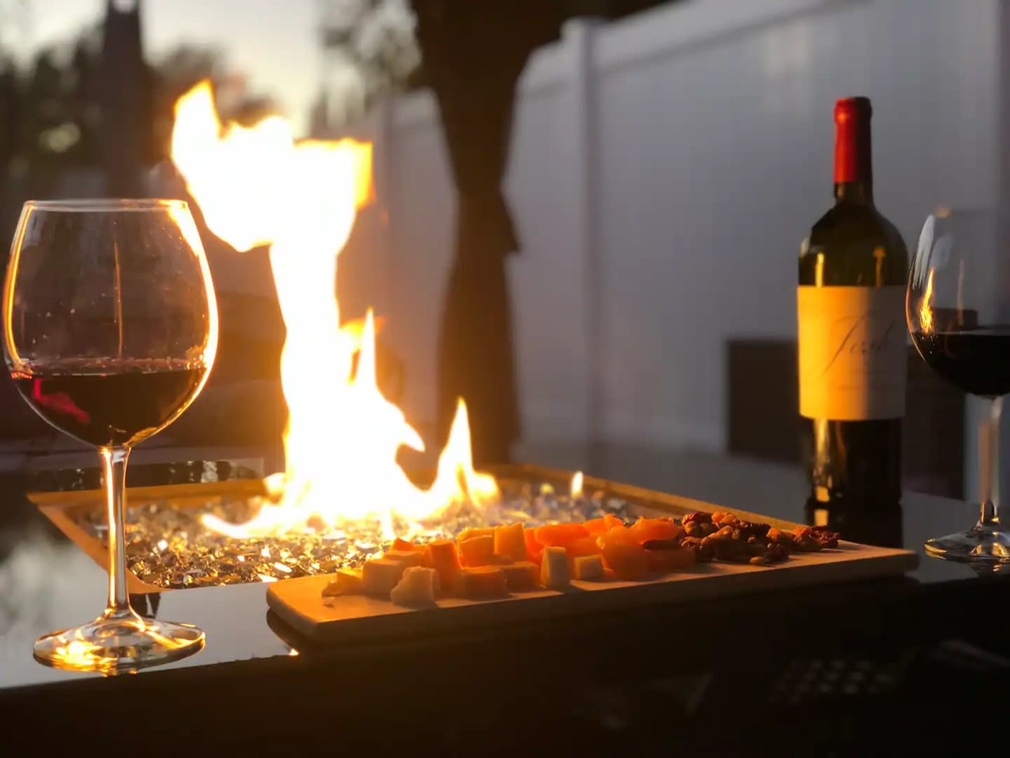 A table setting features two glasses of red wine, a bottle of wine, and a charcuterie board beside a fire pit with flames illuminated against a twilight background.