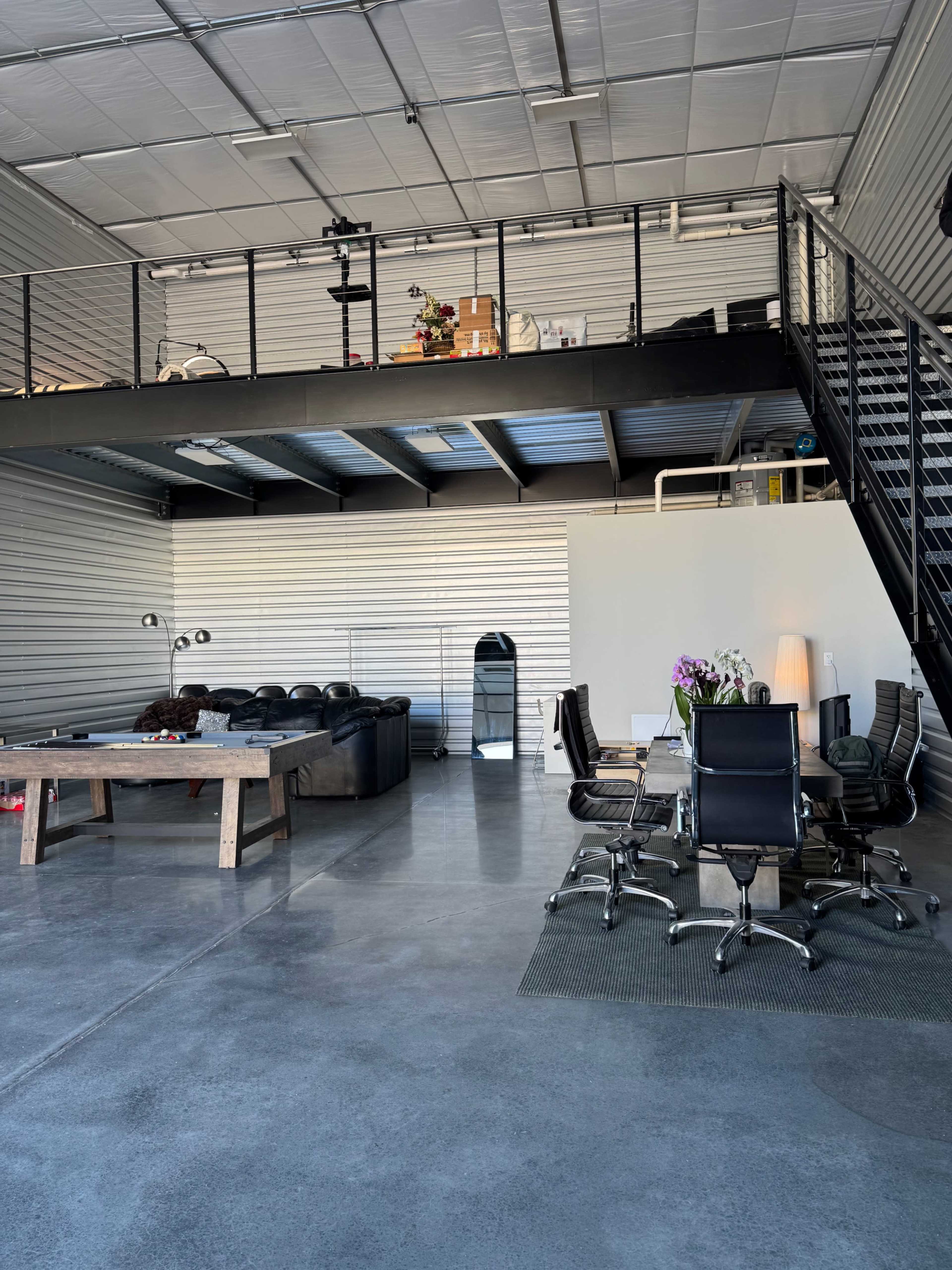 The image shows a spacious industrial-style interior with a lofted area, featuring a seating area with a leather couch and a meeting space with office chairs around a table.
