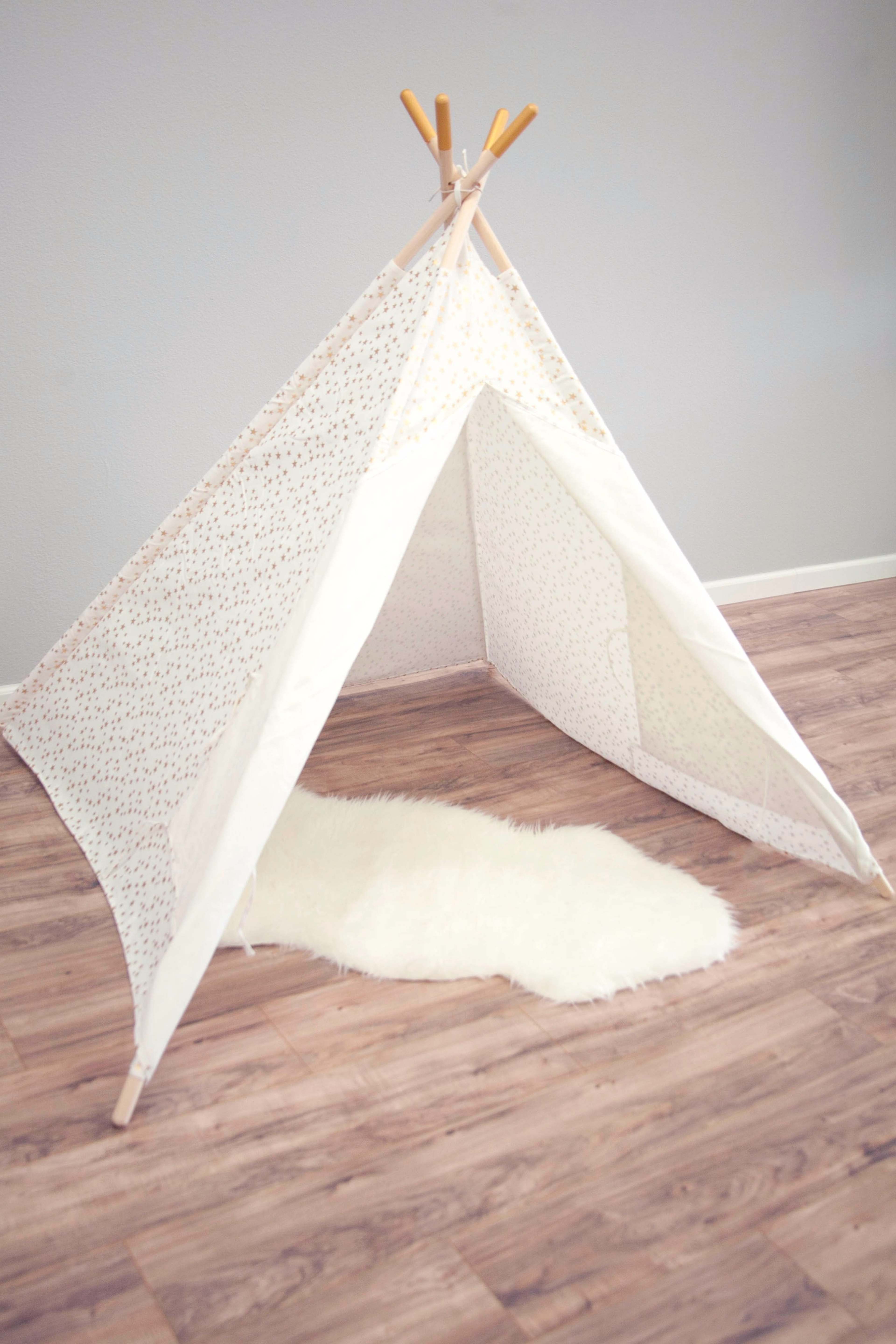 A white fabric teepee stands on a wooden floor, with a soft, white rug placed inside.