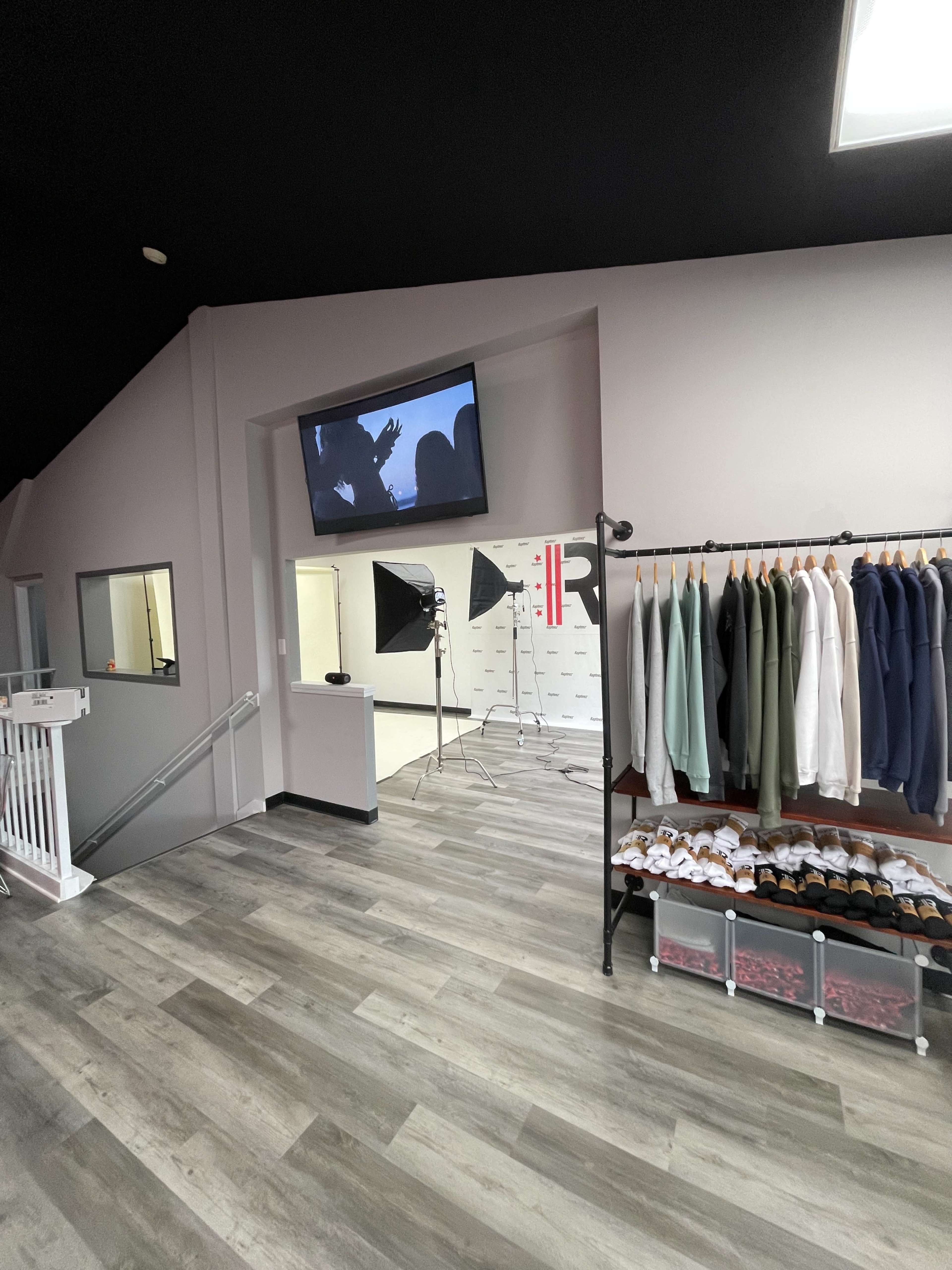 An interior space of a retail store featuring a display of clothing on hangers, a video screen on the wall, and two photography lights set up for a shoot.