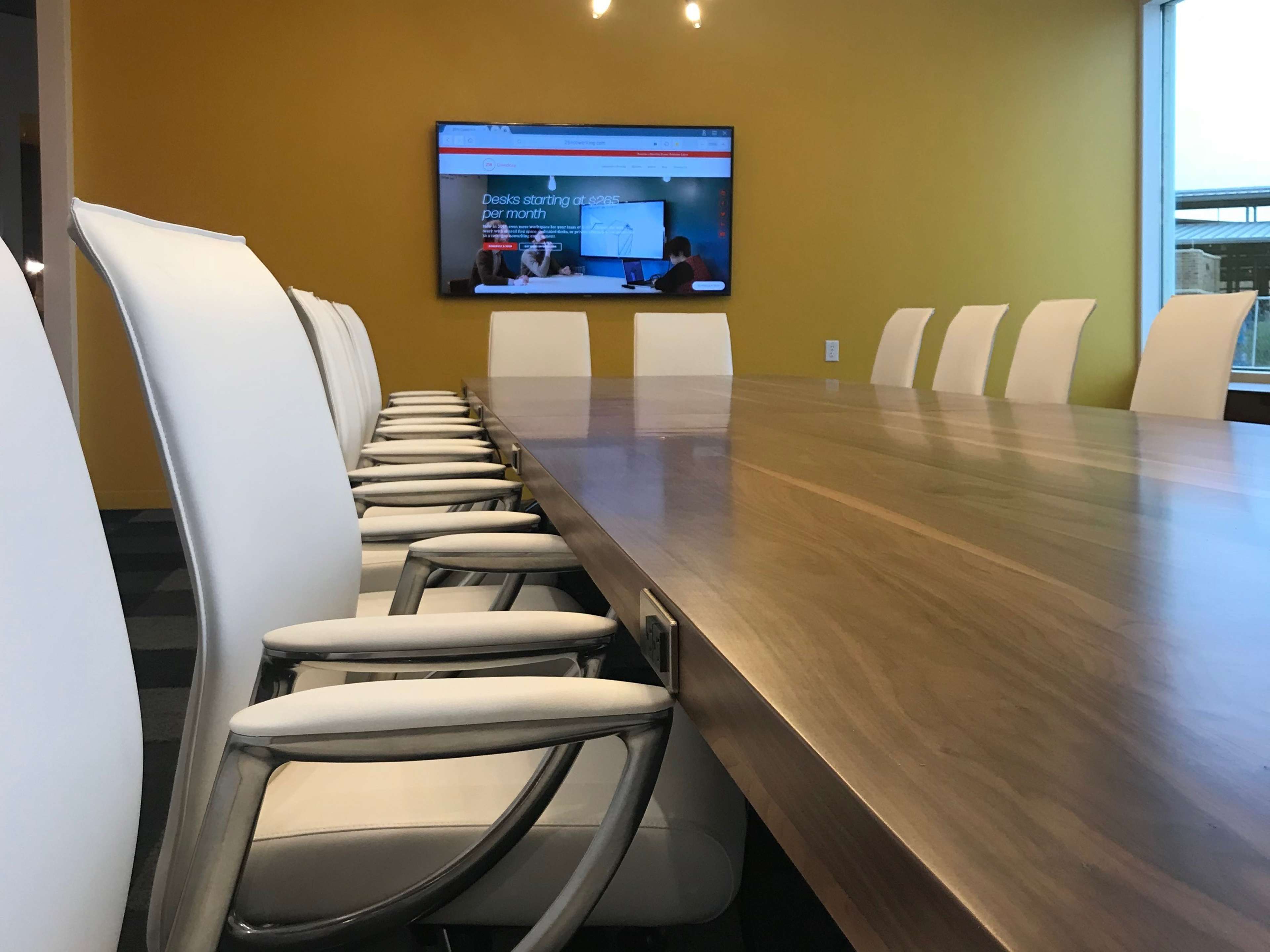 A long wooden conference table is set with white chairs on either side, and a large television screen is mounted on a yellow wall at the far end of the room.