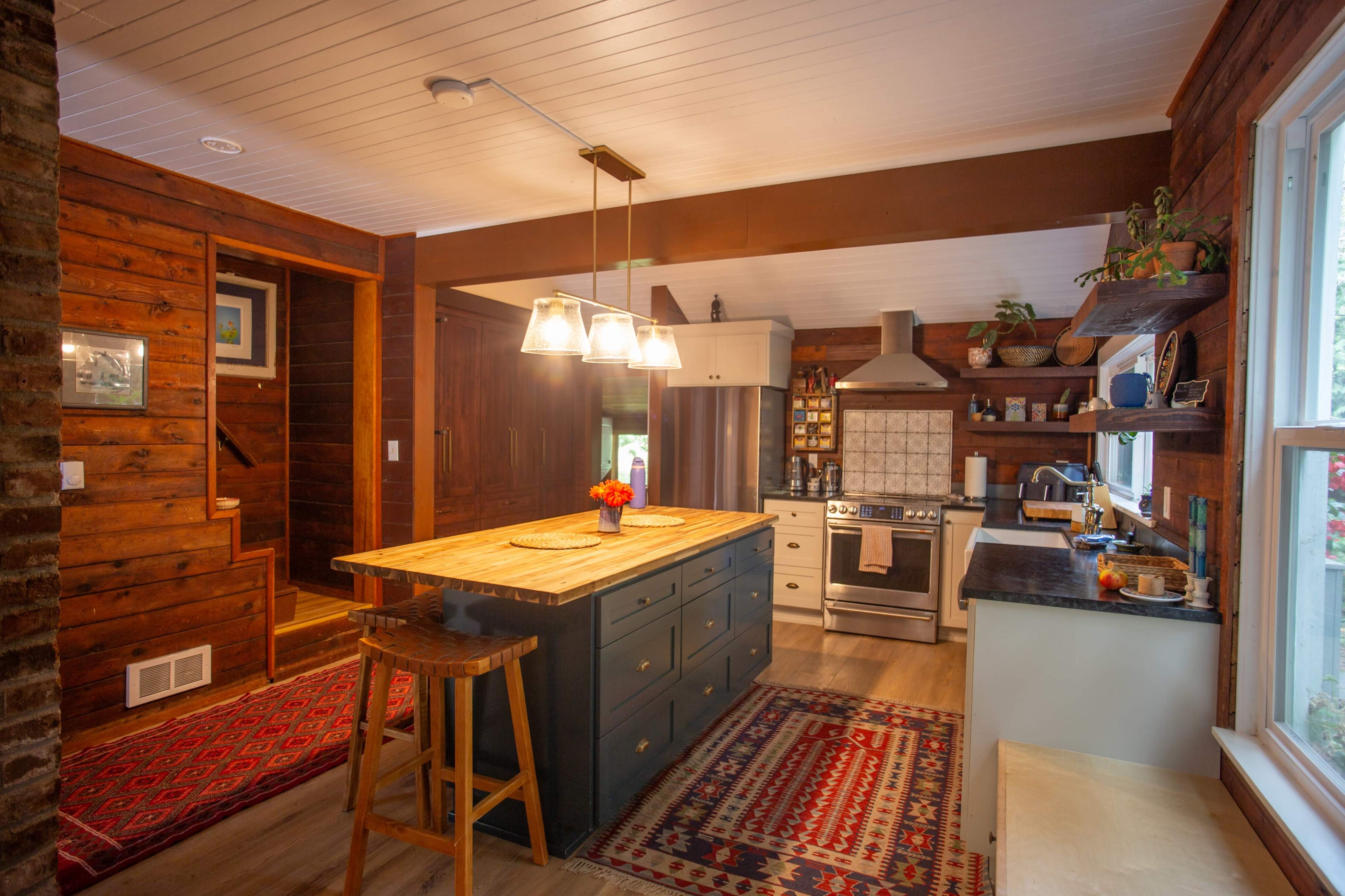 The kitchen features a central island with a wooden countertop, stainless steel appliances, and a blend of dark wood cabinets and open shelving.
