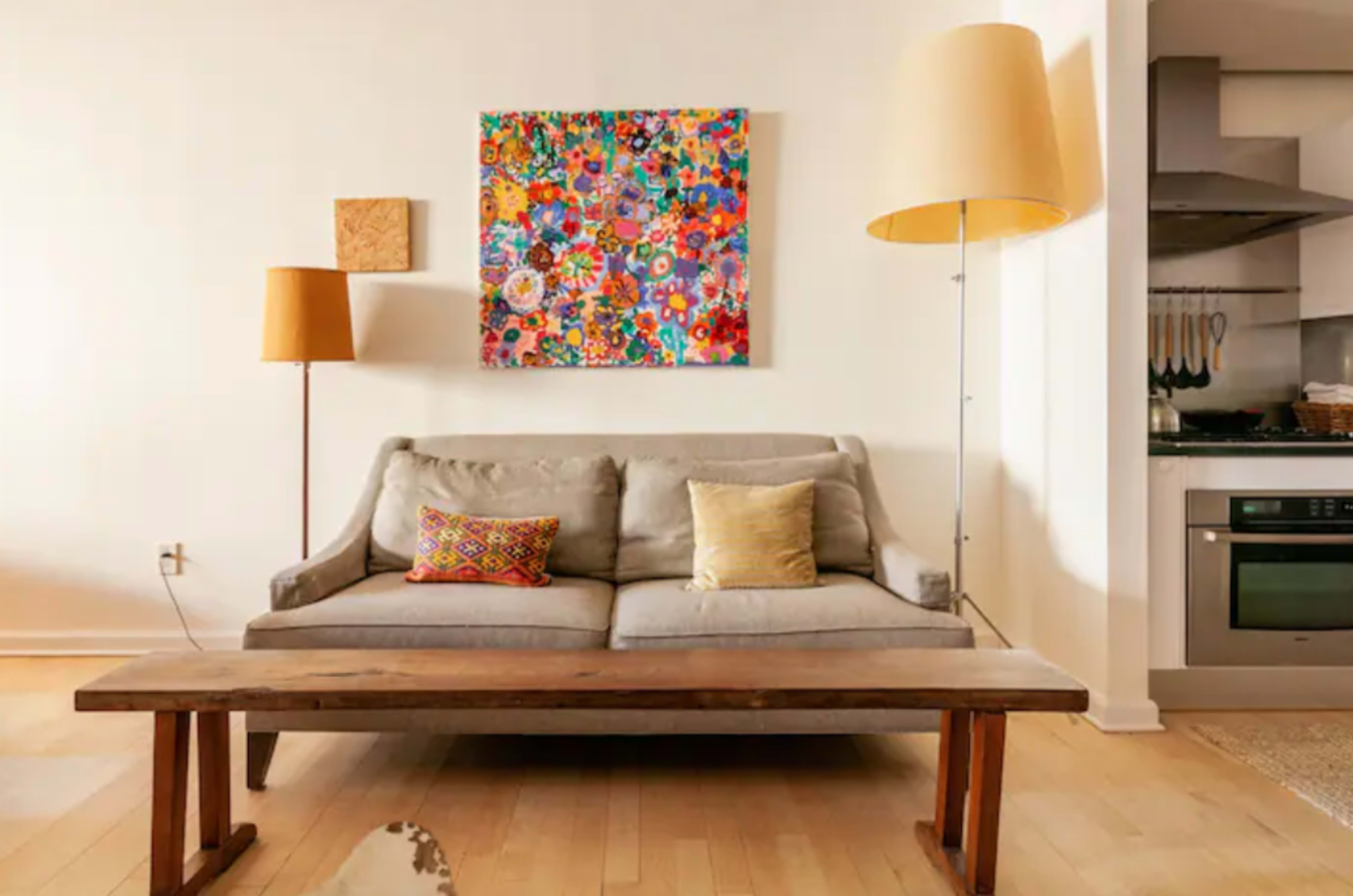 A gray sofa with decorative pillows is positioned in front of a wooden coffee table, with a colorful artwork hanging on the wall and two floor lamps nearby.