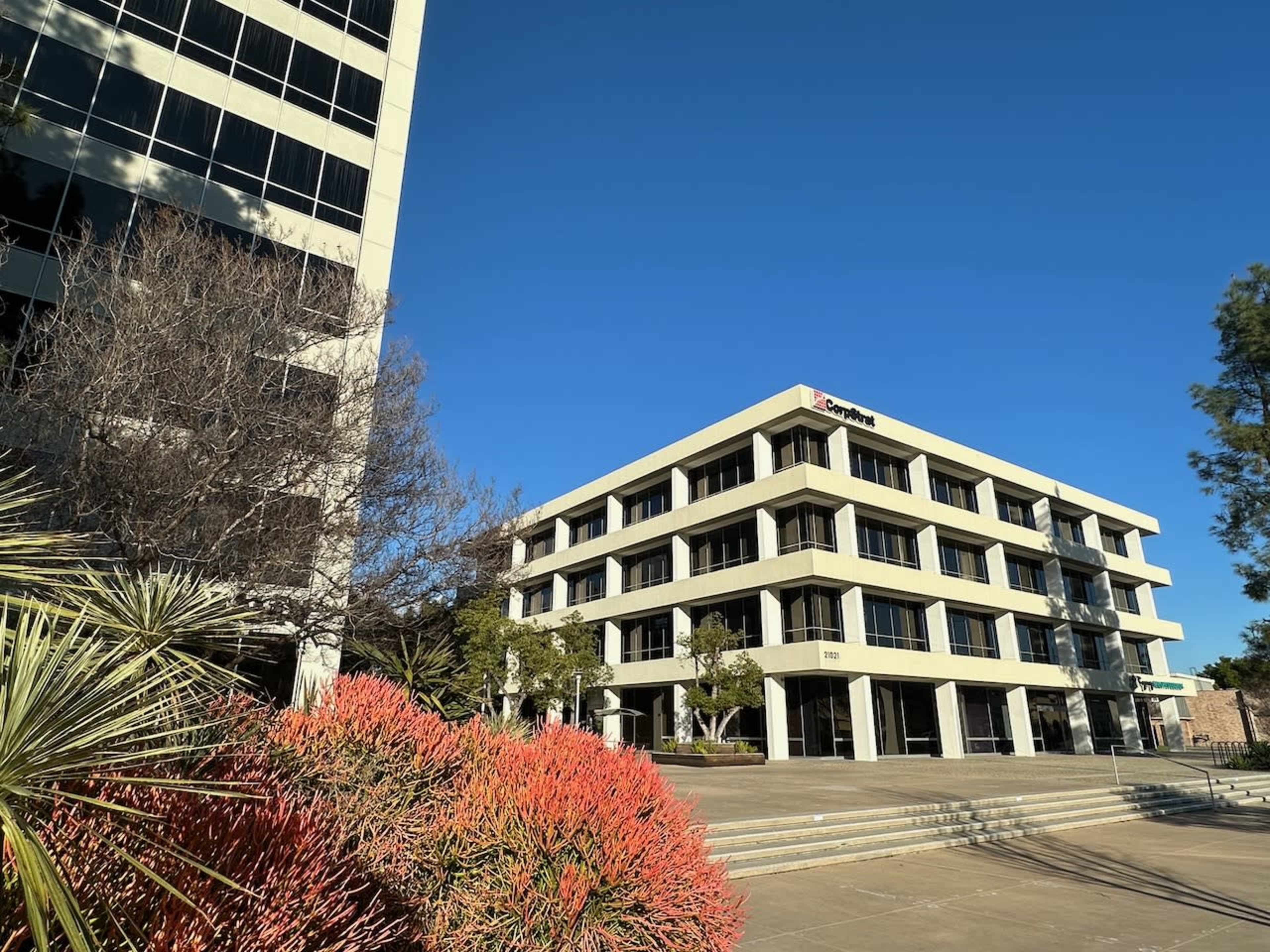 A modern office building is situated under a clear blue sky, surrounded by landscaped greenery and colorful plants.