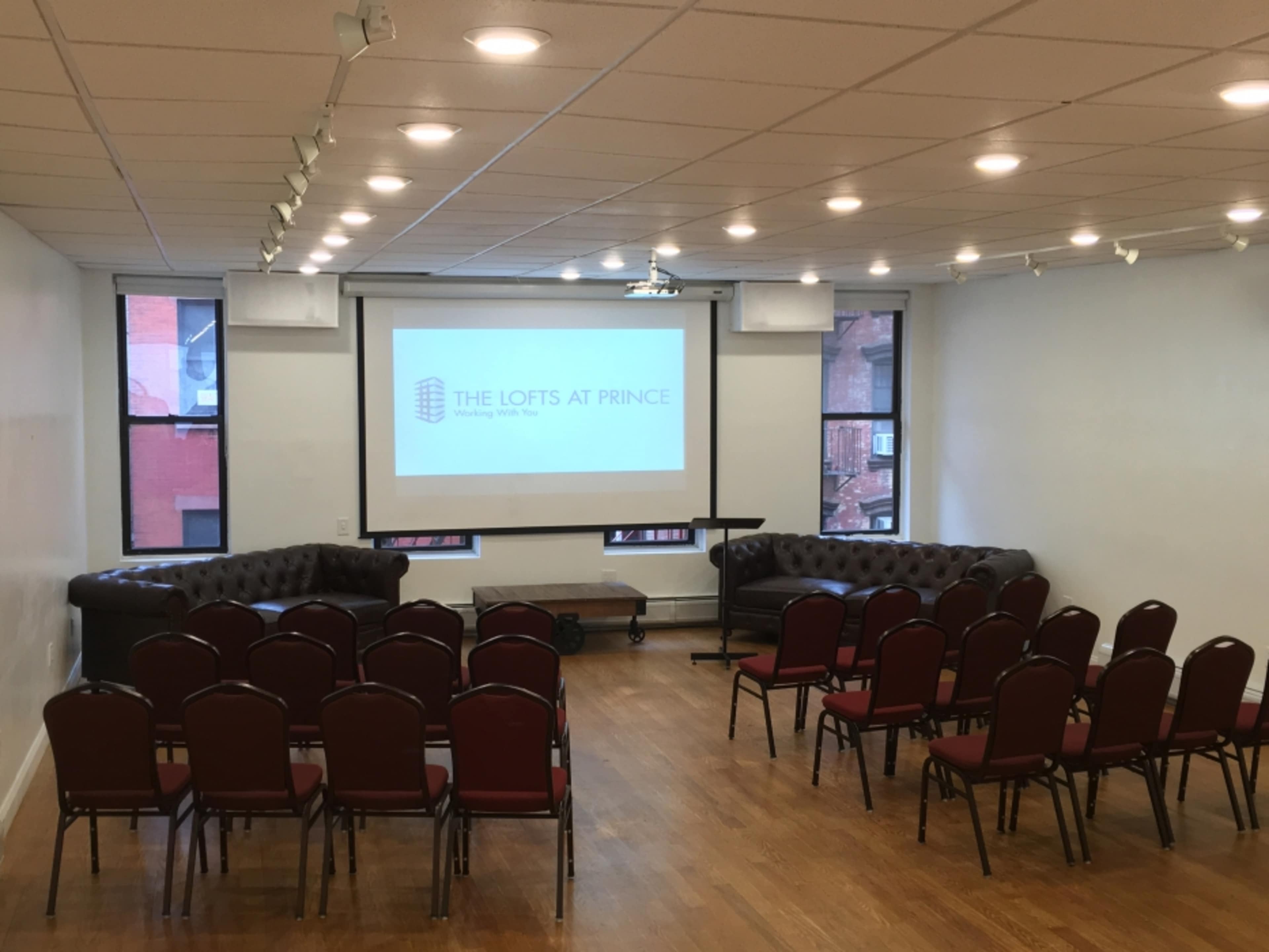 A room is set up for a presentation, featuring rows of red chairs facing a screen with the text "THE LOFTS AT PRINCE."