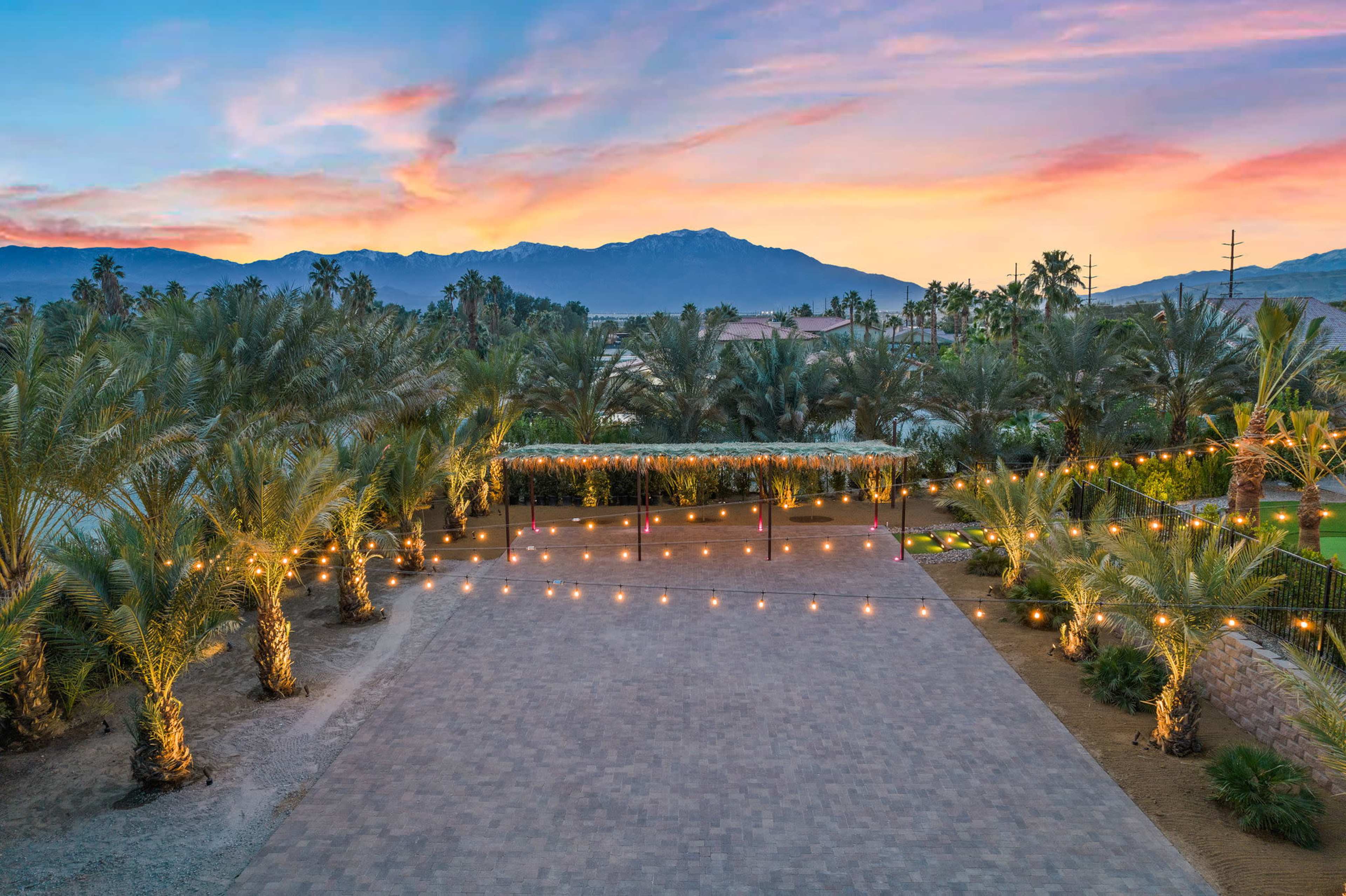 A landscaped area with palm trees and string lights under a colorful sunset, framed by mountains in the background.