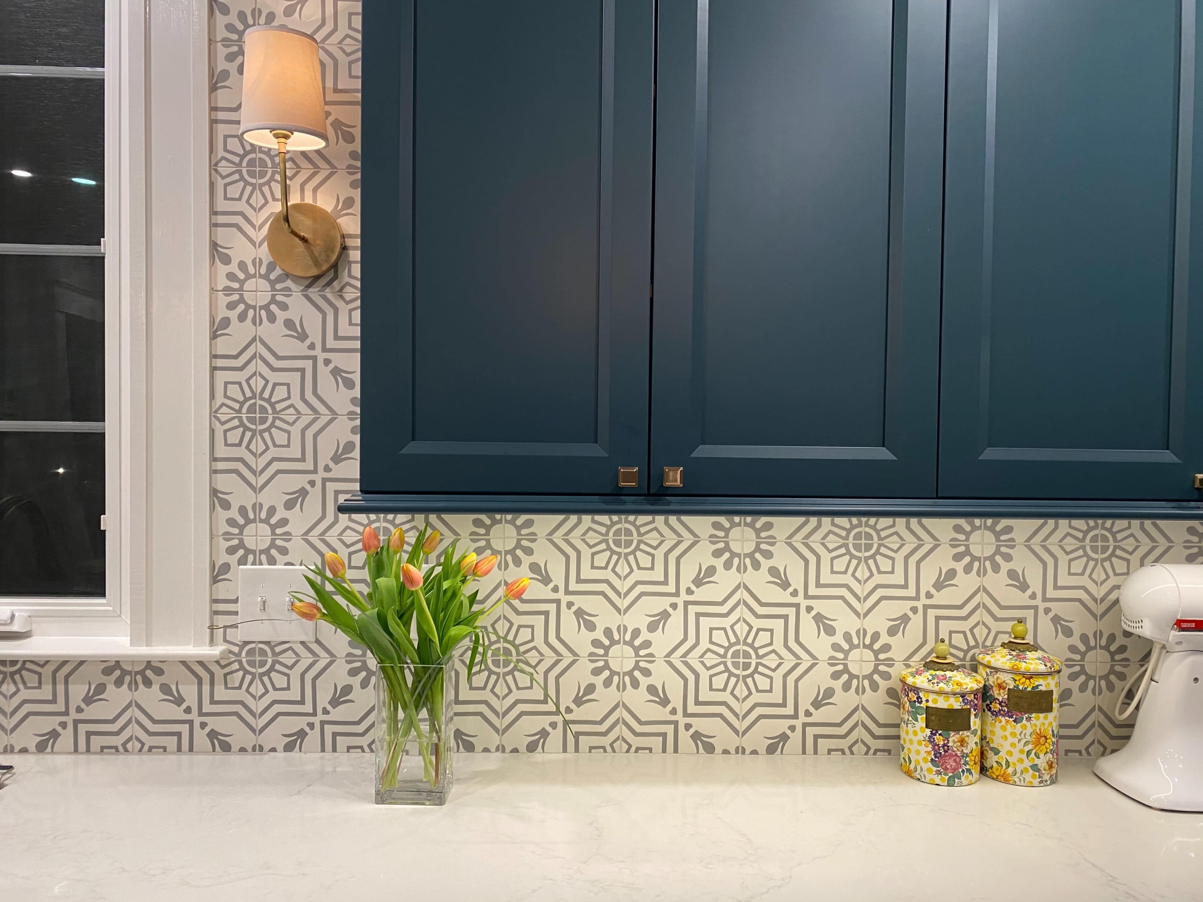 The image shows a kitchen countertop with a vase of tulips, decorative canisters, and patterned backsplash behind dark blue cabinets.