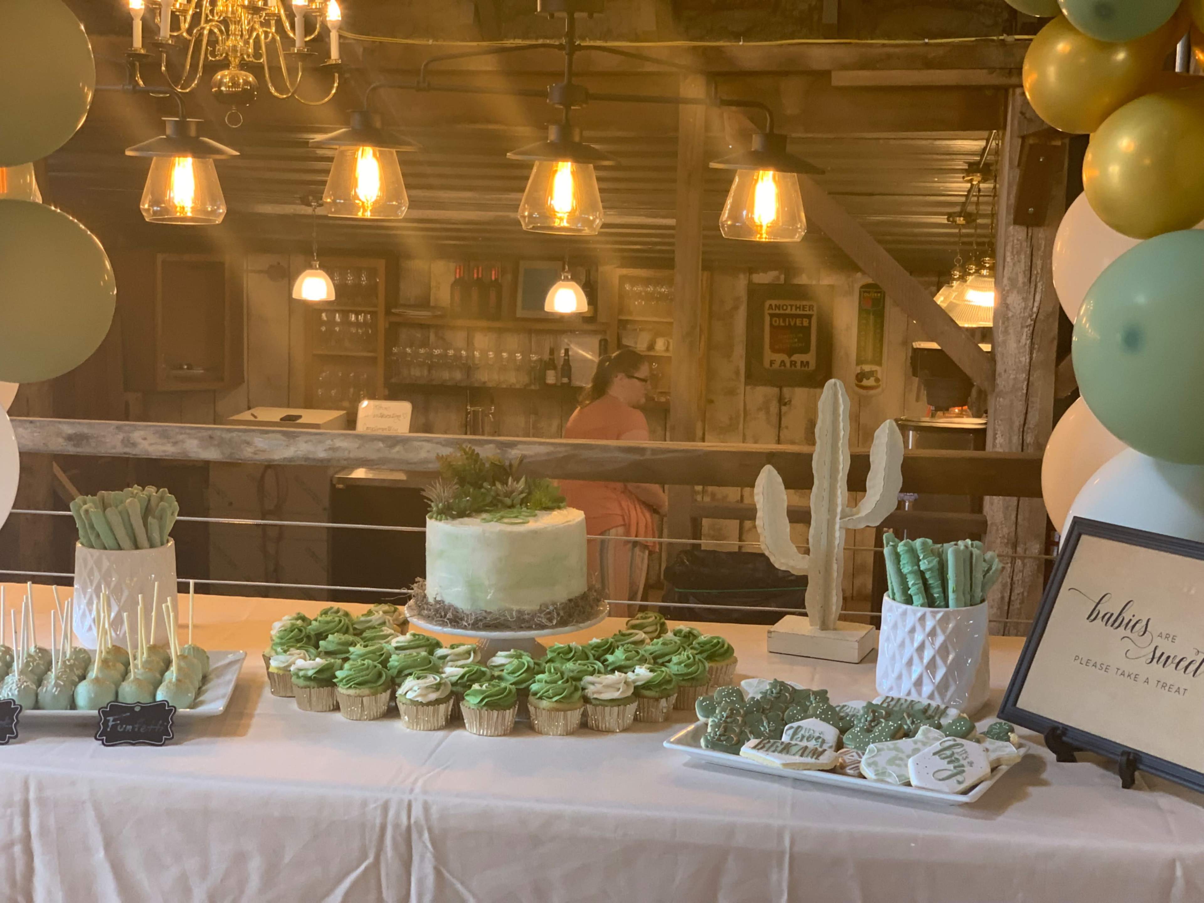 A dessert table is set up with a cake, cupcakes, and various treats, decorated with green icing, next to a bar in a rustic venue with warm lighting.