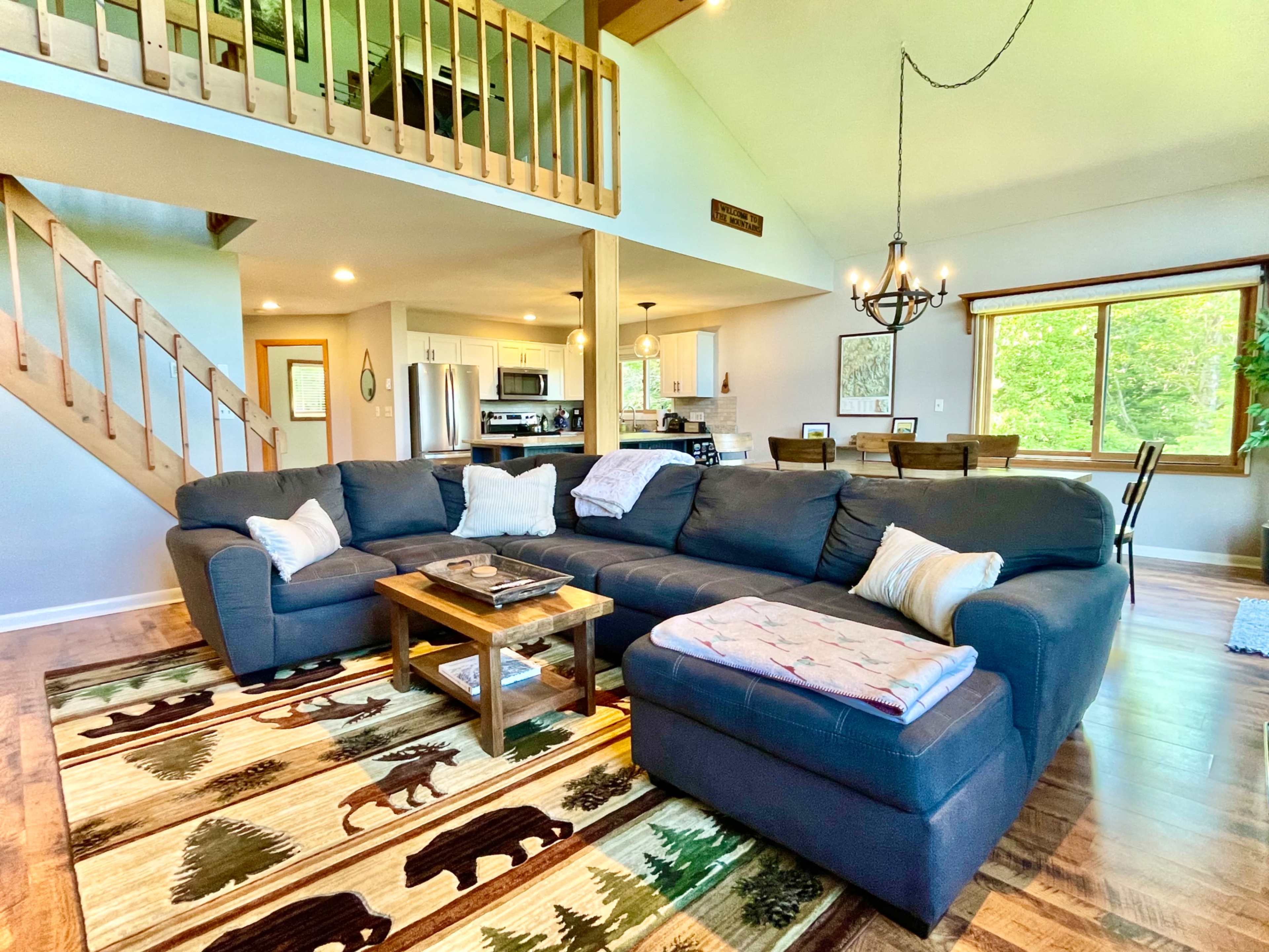 The image shows a cozy living room with a large sectional sofa, a coffee table, and a decorative rug featuring bears and trees, adjacent to a dining area and kitchen.
