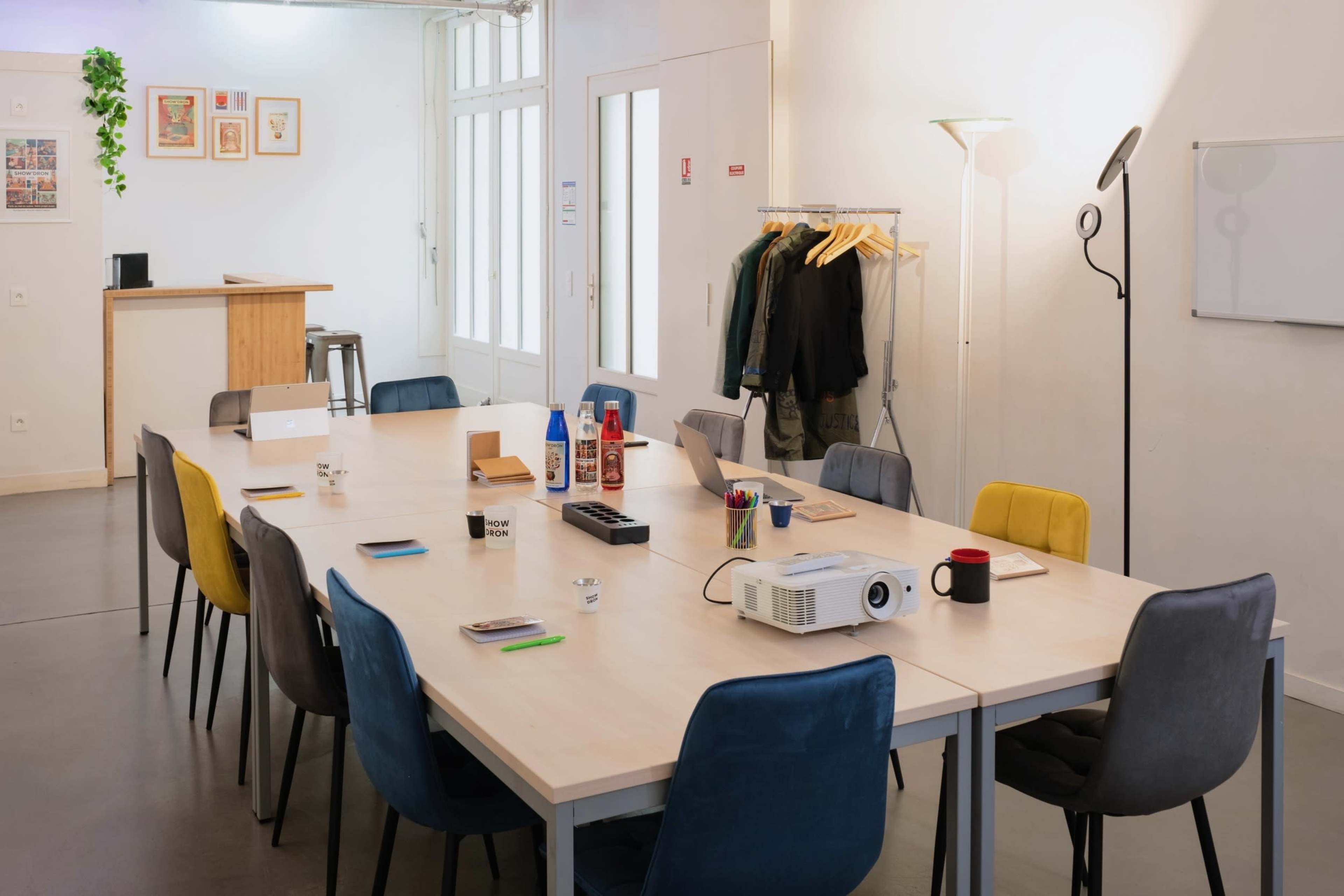 The image shows a modern meeting room with a long wooden table surrounded by colorful chairs, a projector, and various office supplies arranged neatly on the table.