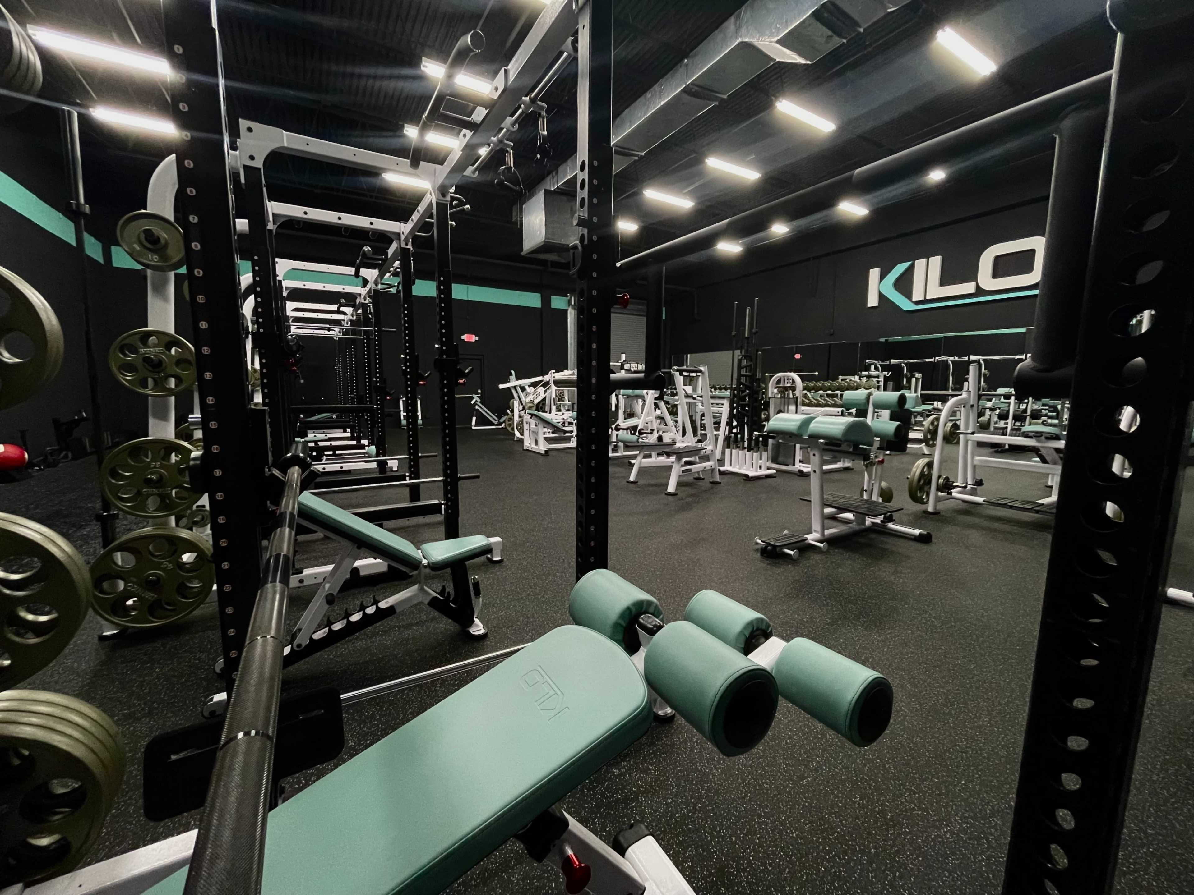 A well-equipped gym features various weightlifting machines and benches under bright overhead lighting.