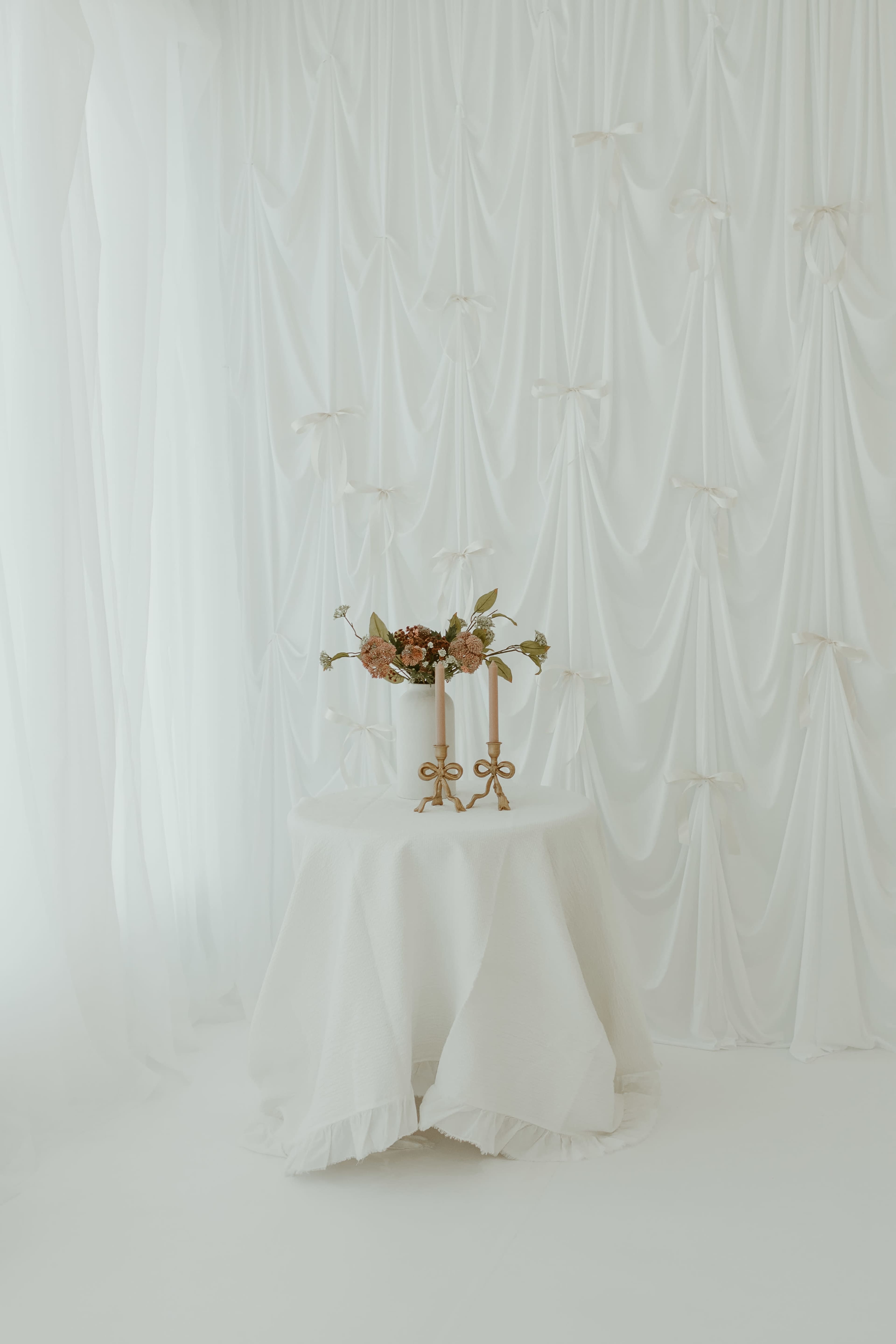 A small round table covered with a white tablecloth holds a floral arrangement and two candle holders in front of a backdrop of draped white fabric.