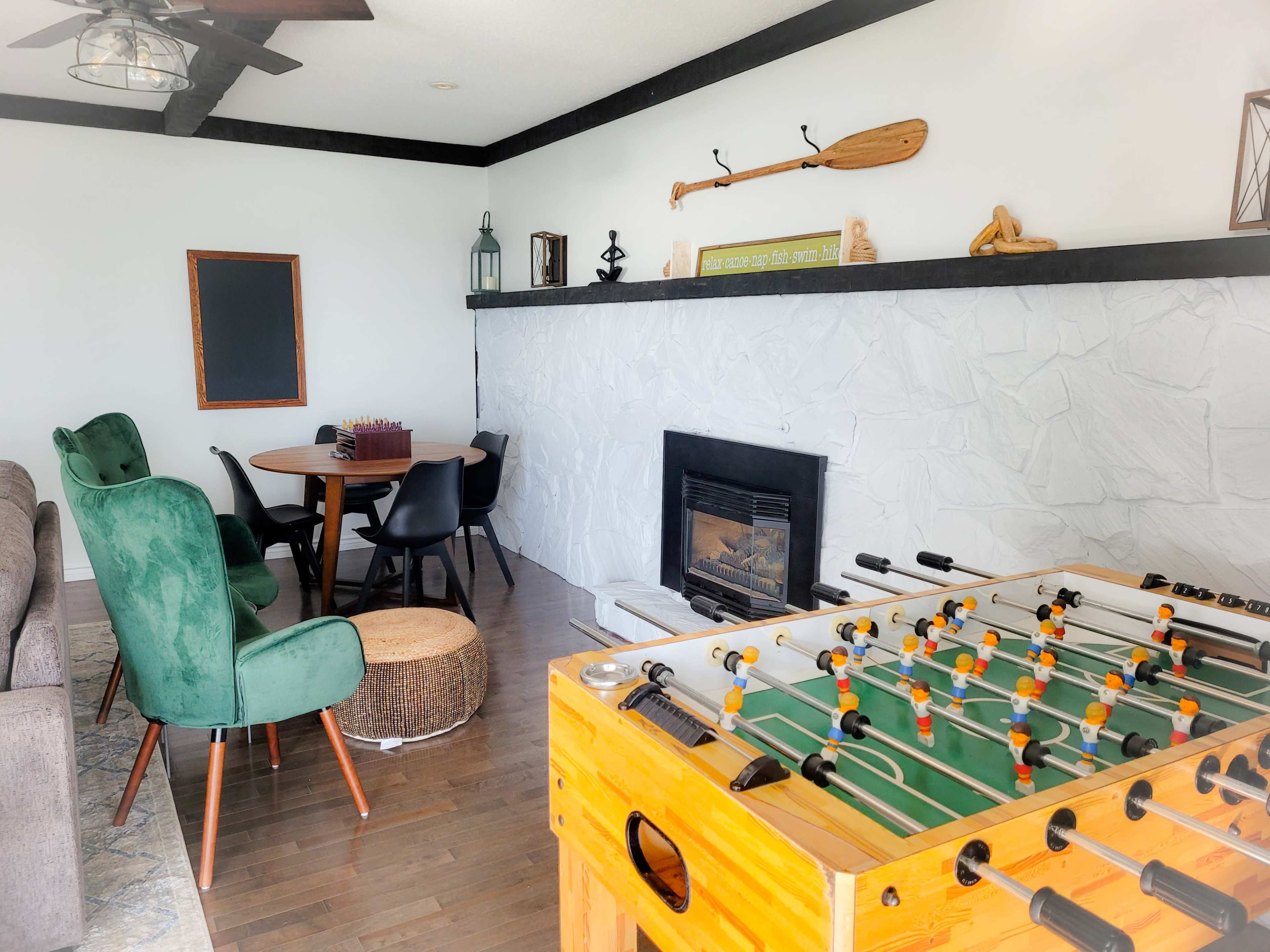 A cozy living area with a foosball table in the foreground, a round dining table, and a stone fireplace against a white wall.