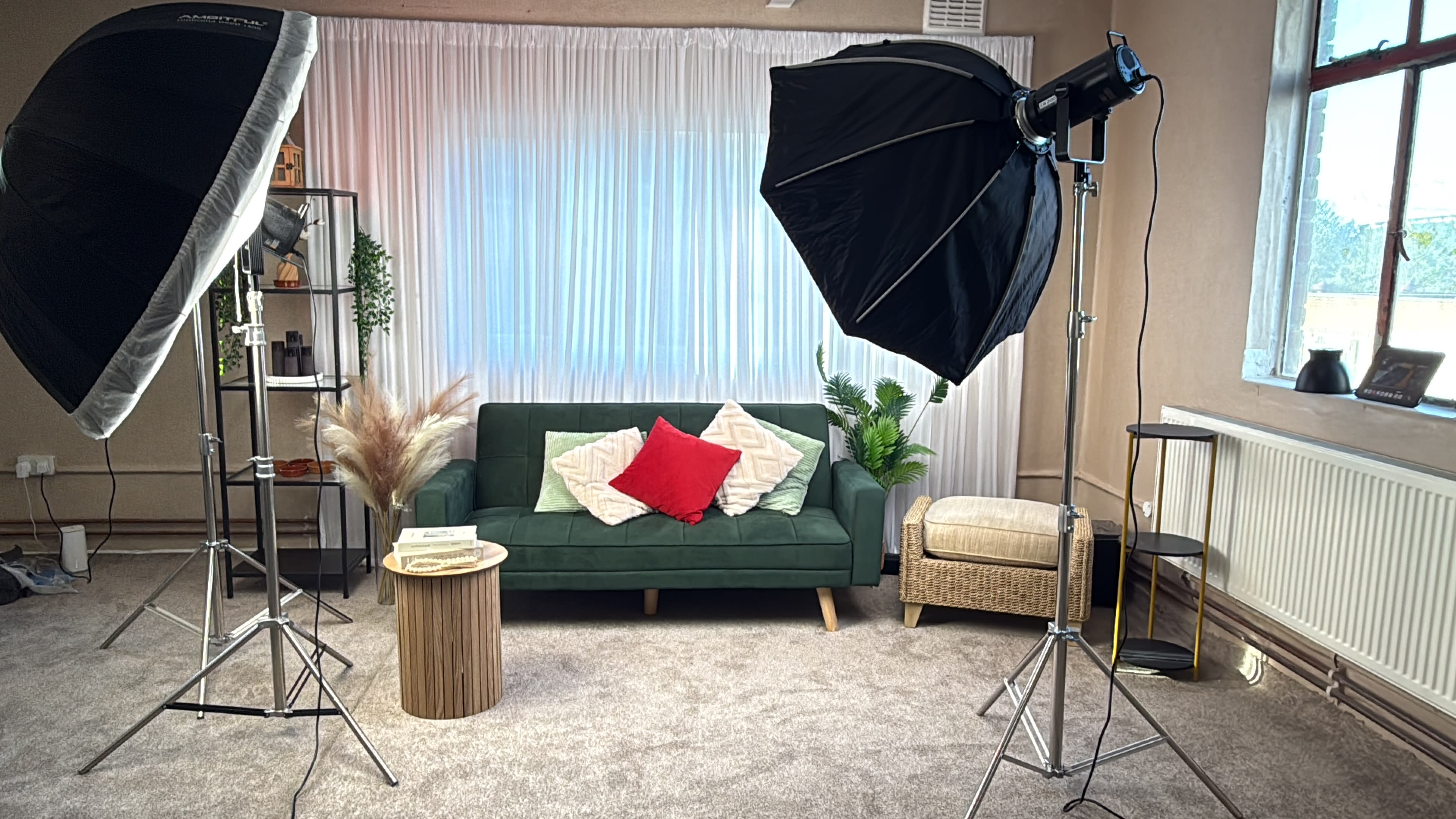 The image shows a cozy living room setup with a green sofa adorned with red and white cushions, surrounded by photography lighting equipment and decorative plants.