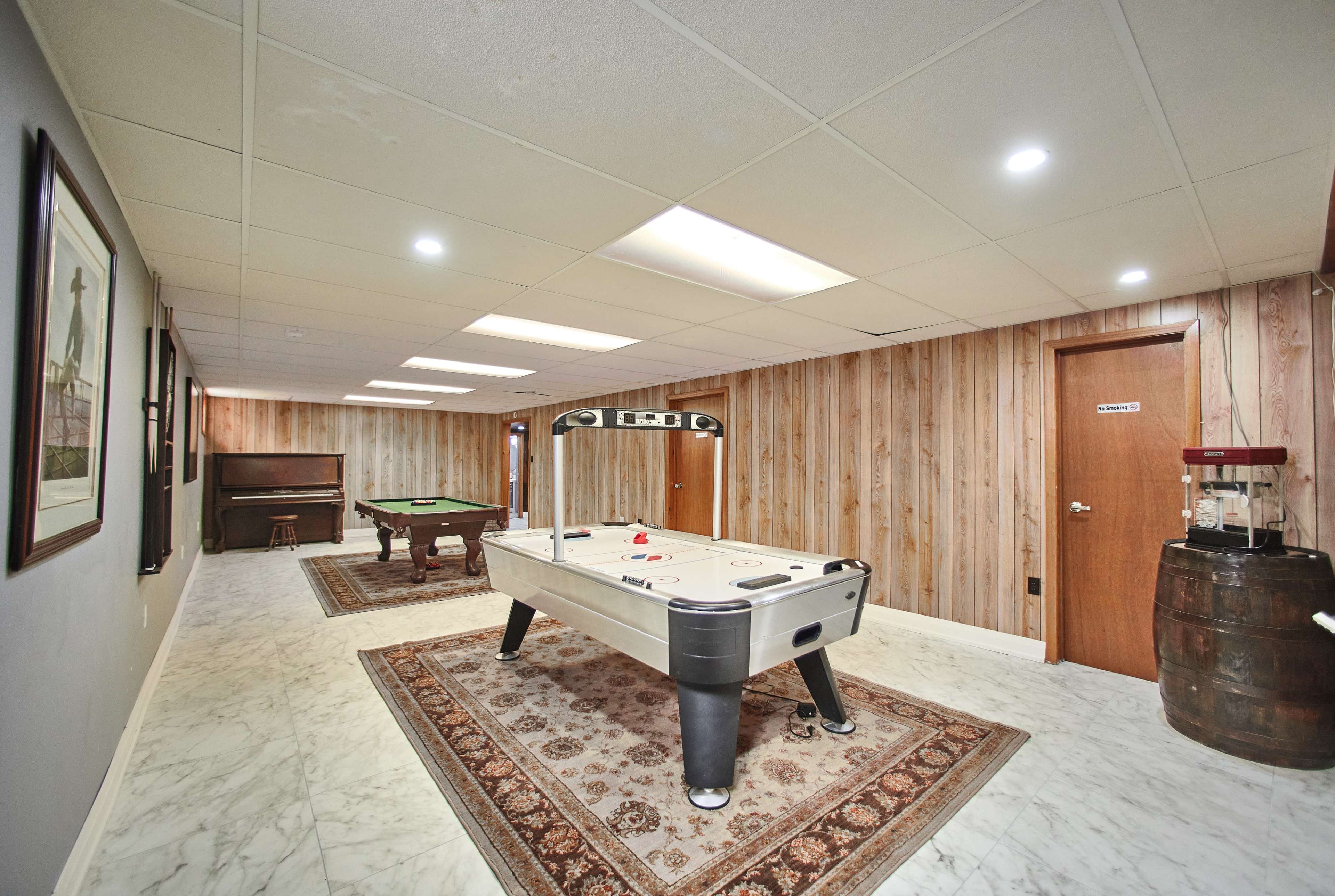 A spacious game room featuring a pool table, air hockey table, and a piano, with wood-paneled walls and marble flooring.