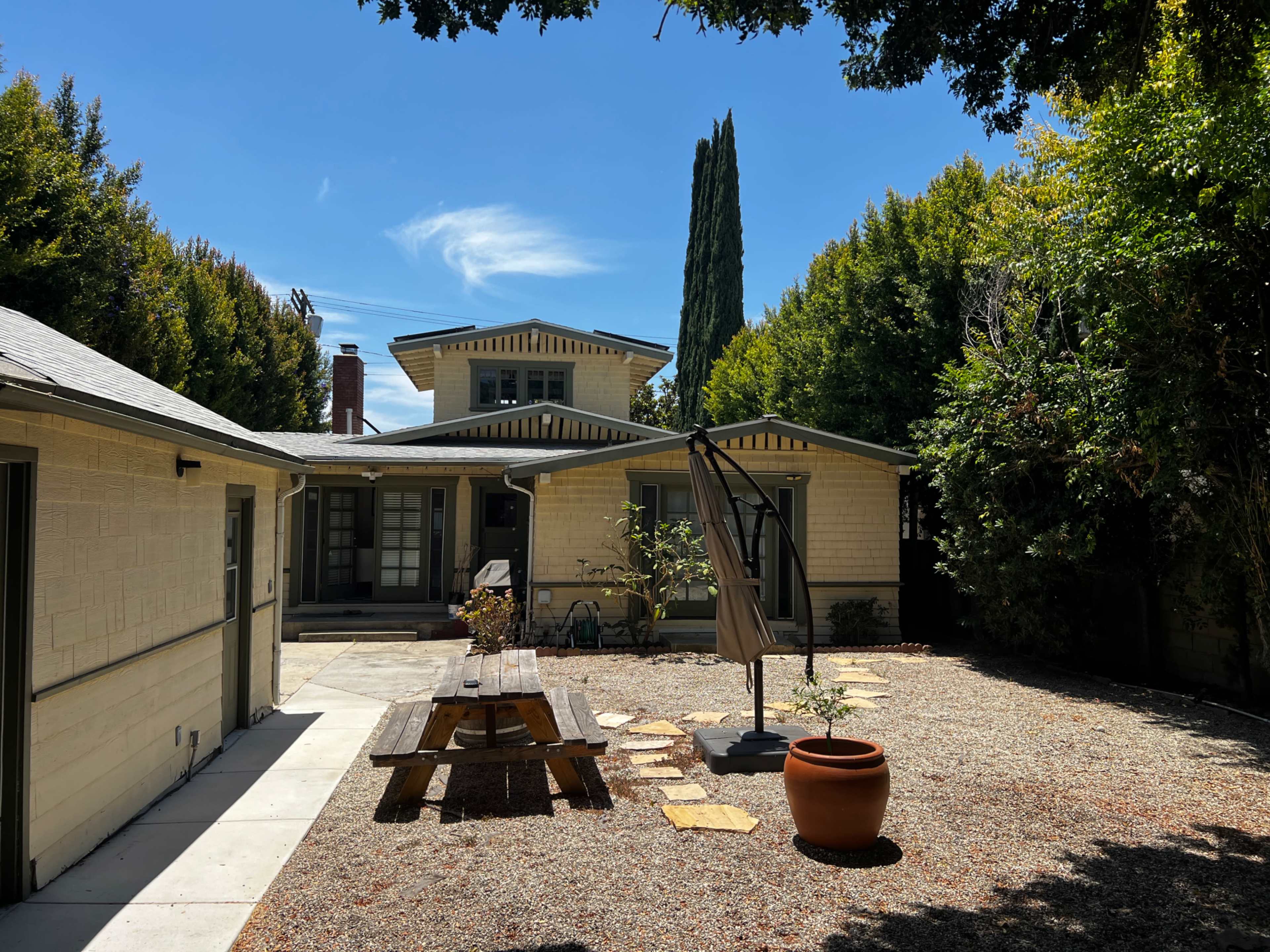 The image shows a residential backyard featuring a picnic table, a large potted plant, and a patio area surrounded by trees and a beige two-story house.
