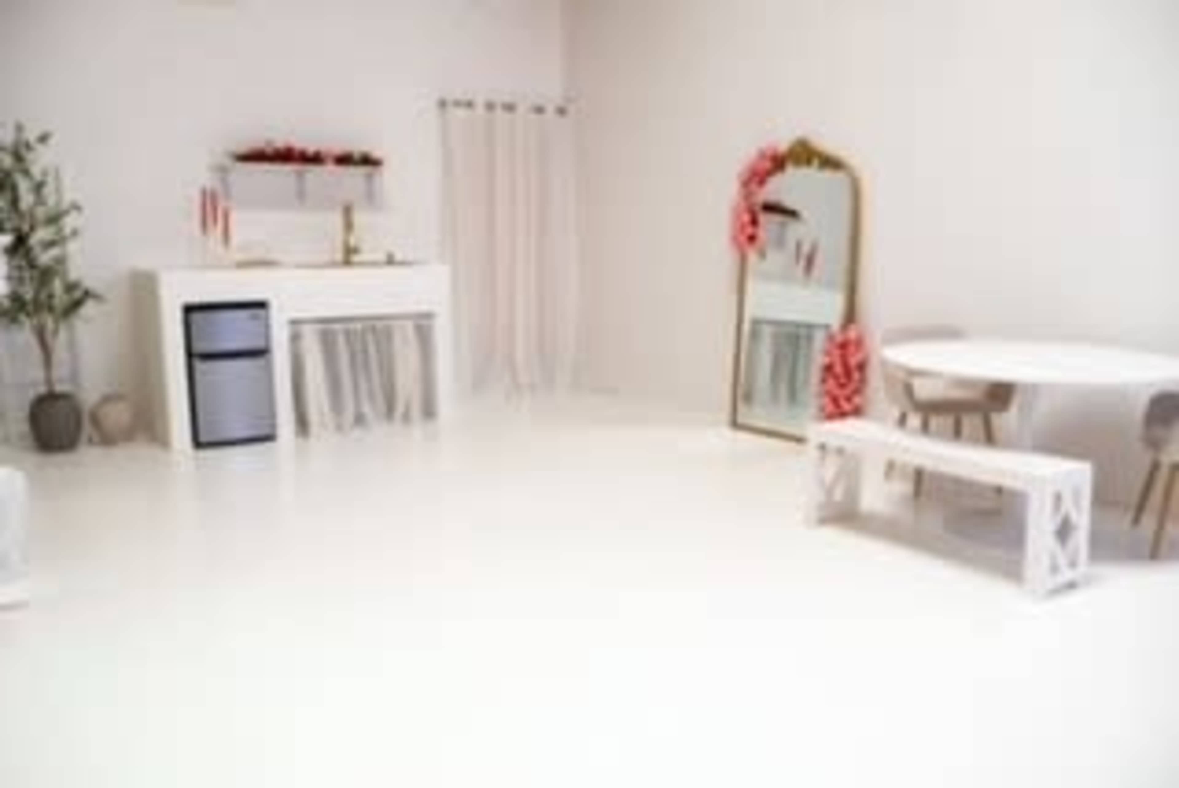 The image shows a bright, minimalist interior space featuring white walls, a white table, a bench, a mirror, and a small kitchenette with a fridge.