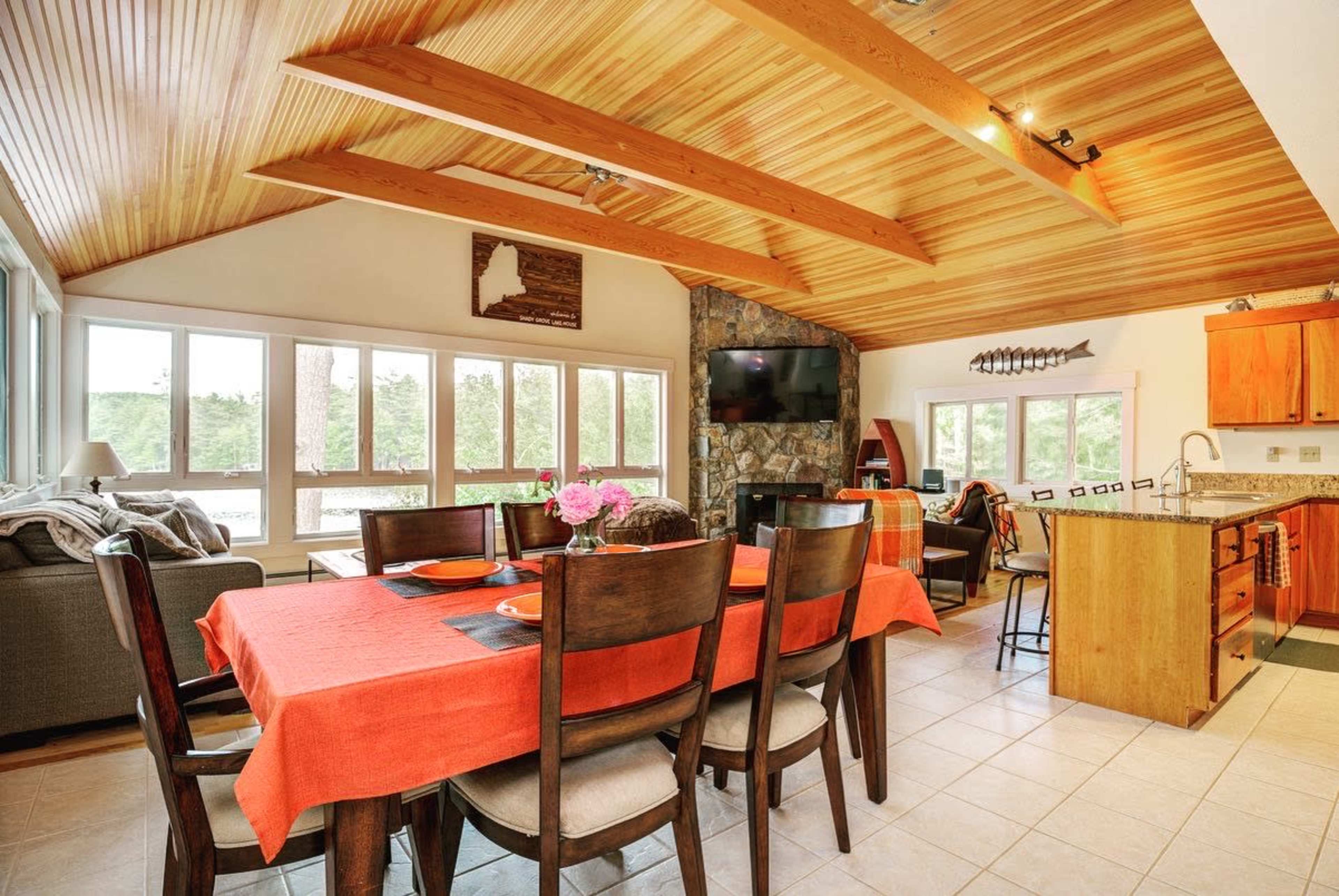 The image shows a bright, open-concept living and dining area with a wooden ceiling, large windows, and a central table set for dining.