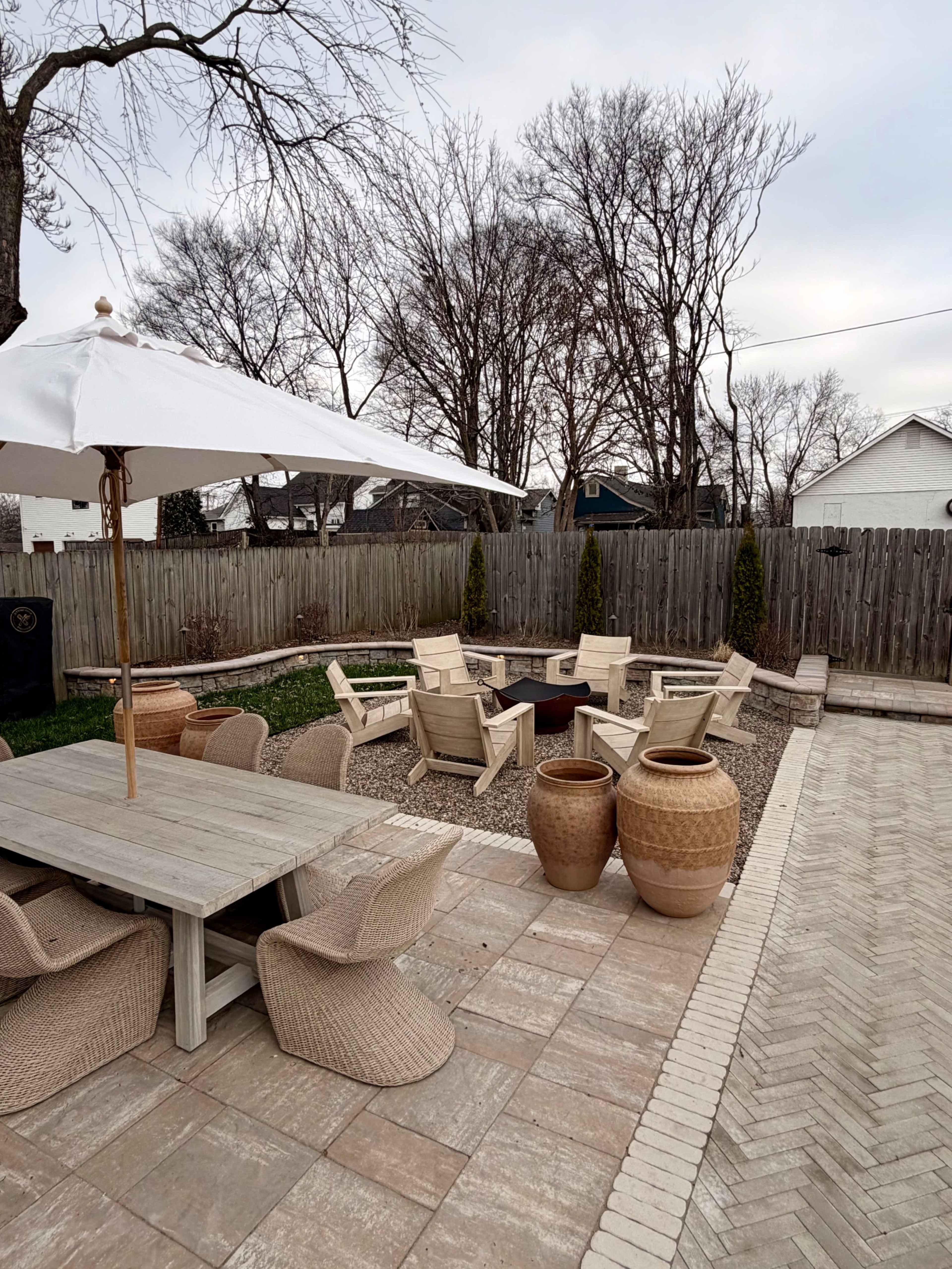 The image shows a patio area with a large table and chairs, surrounded by a gravel space with multiple lounge chairs and two large ceramic pots, set against a backdrop of bare trees and wooden fencing.