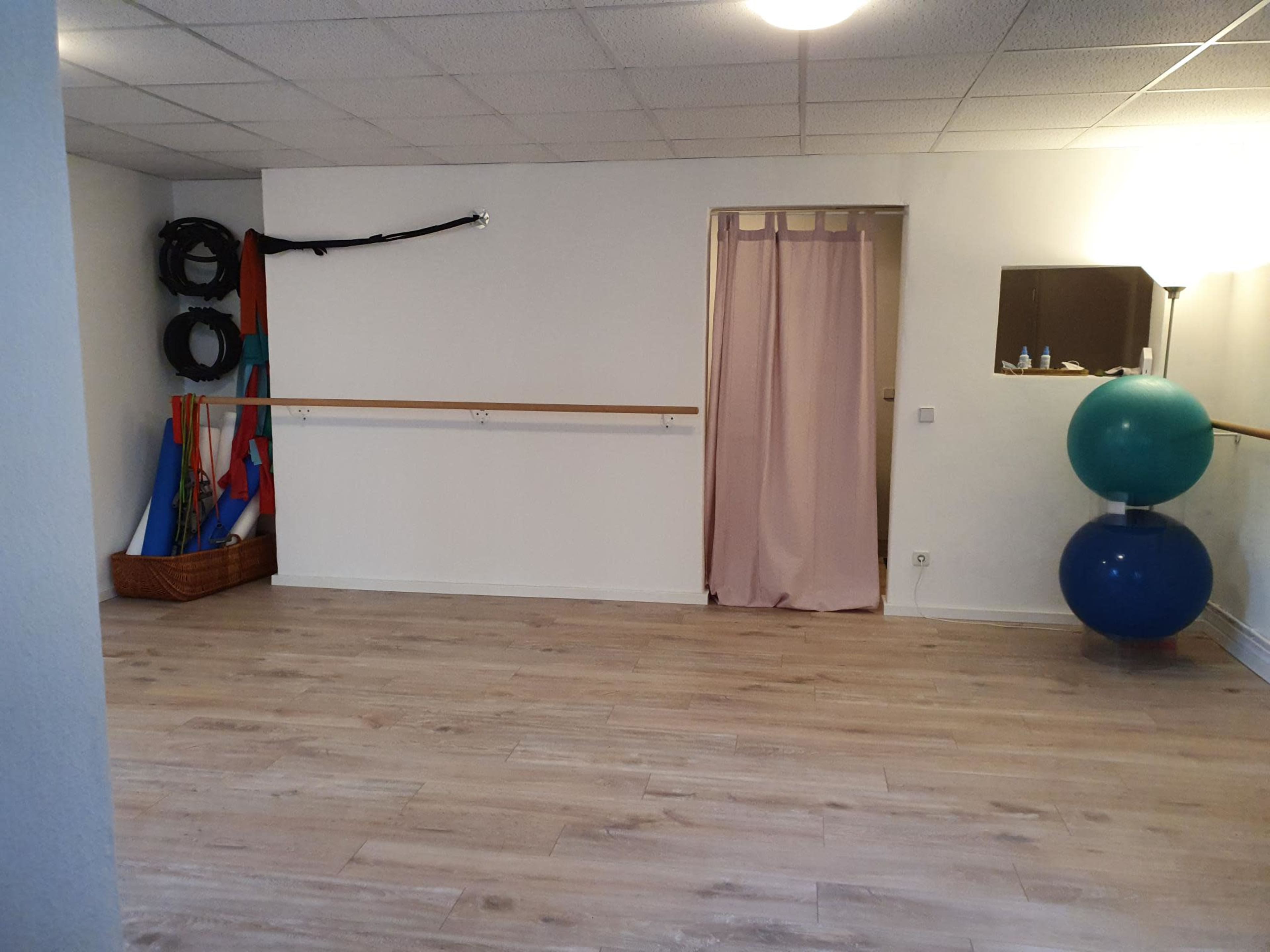 The image shows a spacious, empty room with a wooden floor, exercise equipment stored against the wall, and a curtain leading to another area.
