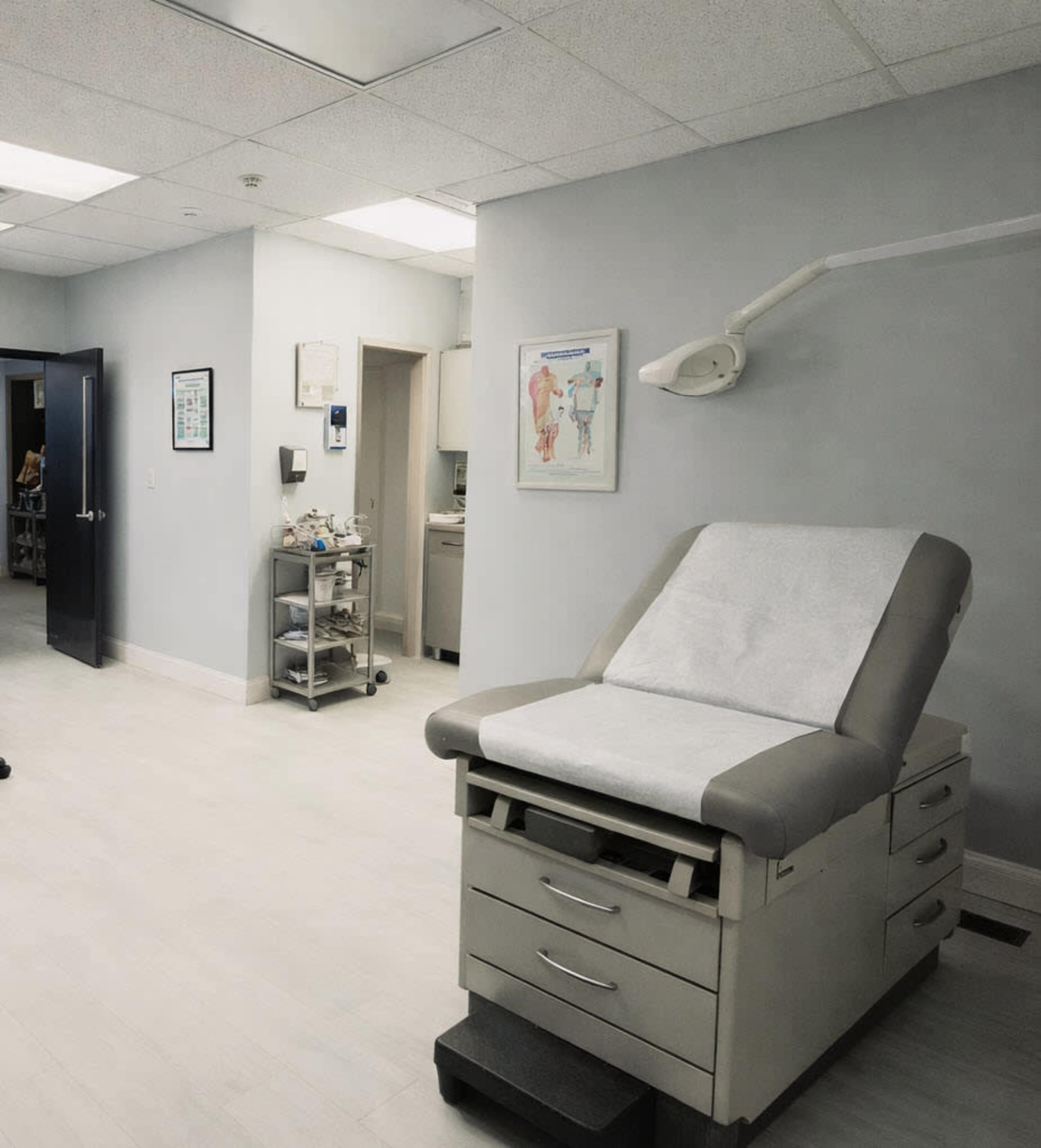 The image shows a medical examination room with an examination table, medical equipment, and a chart on the wall.