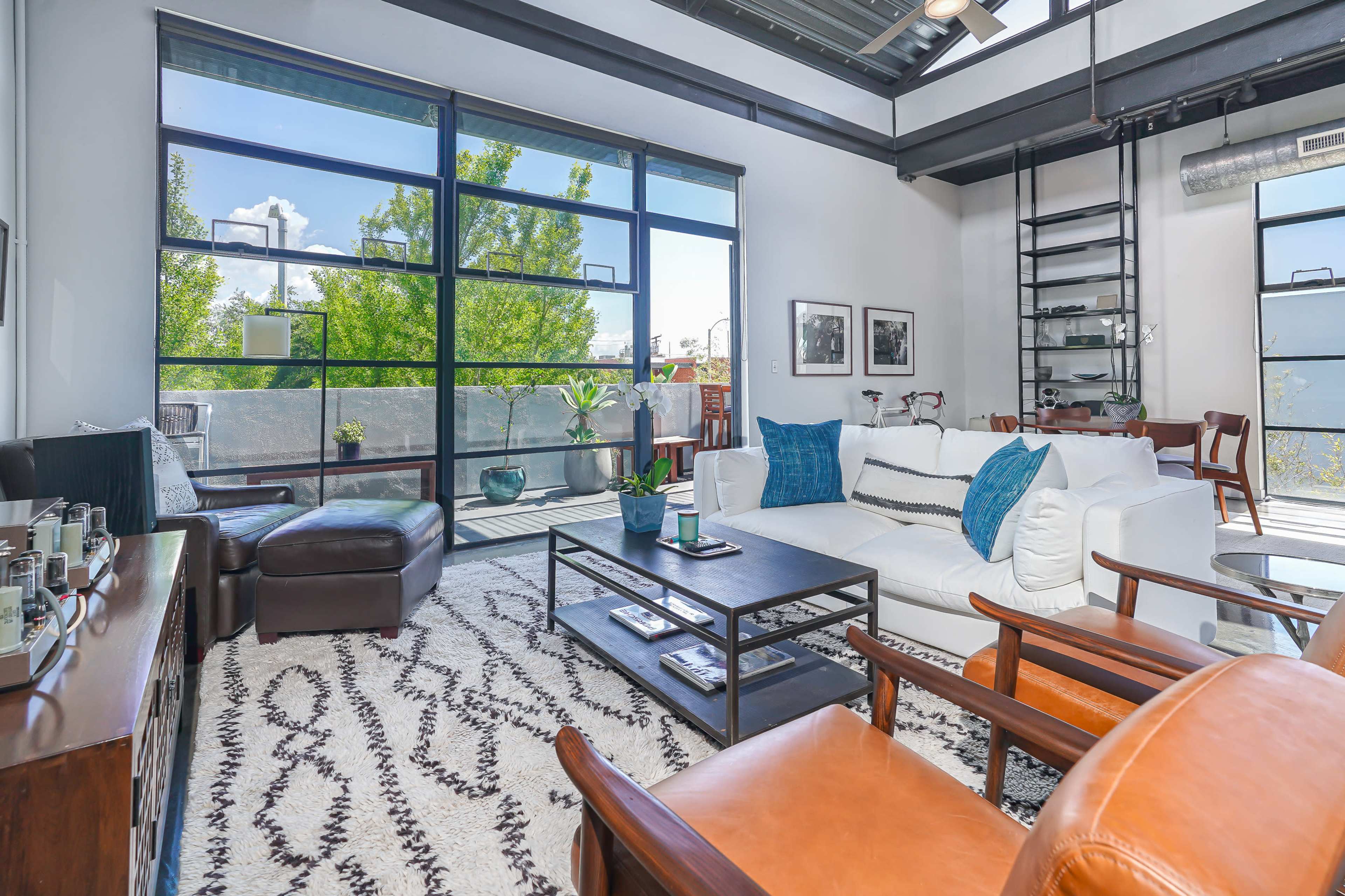 The image shows a modern living room with large windows, a white sofa, two wooden chairs, and various decorative plants.