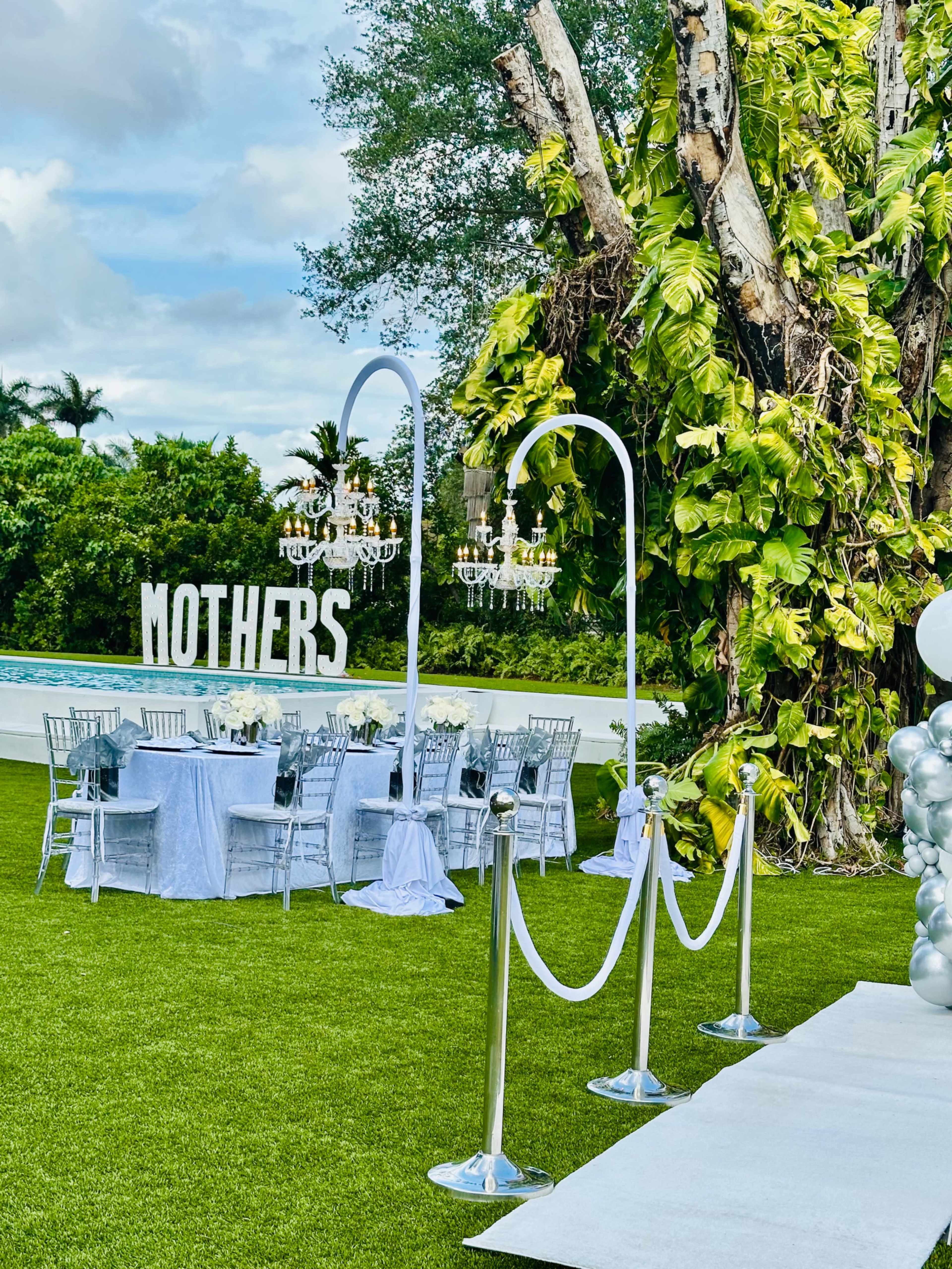 The image shows a garden setup for a Mother's Day event, featuring white seating, decorative chandeliers, and a large "MOTHERS" sign.
