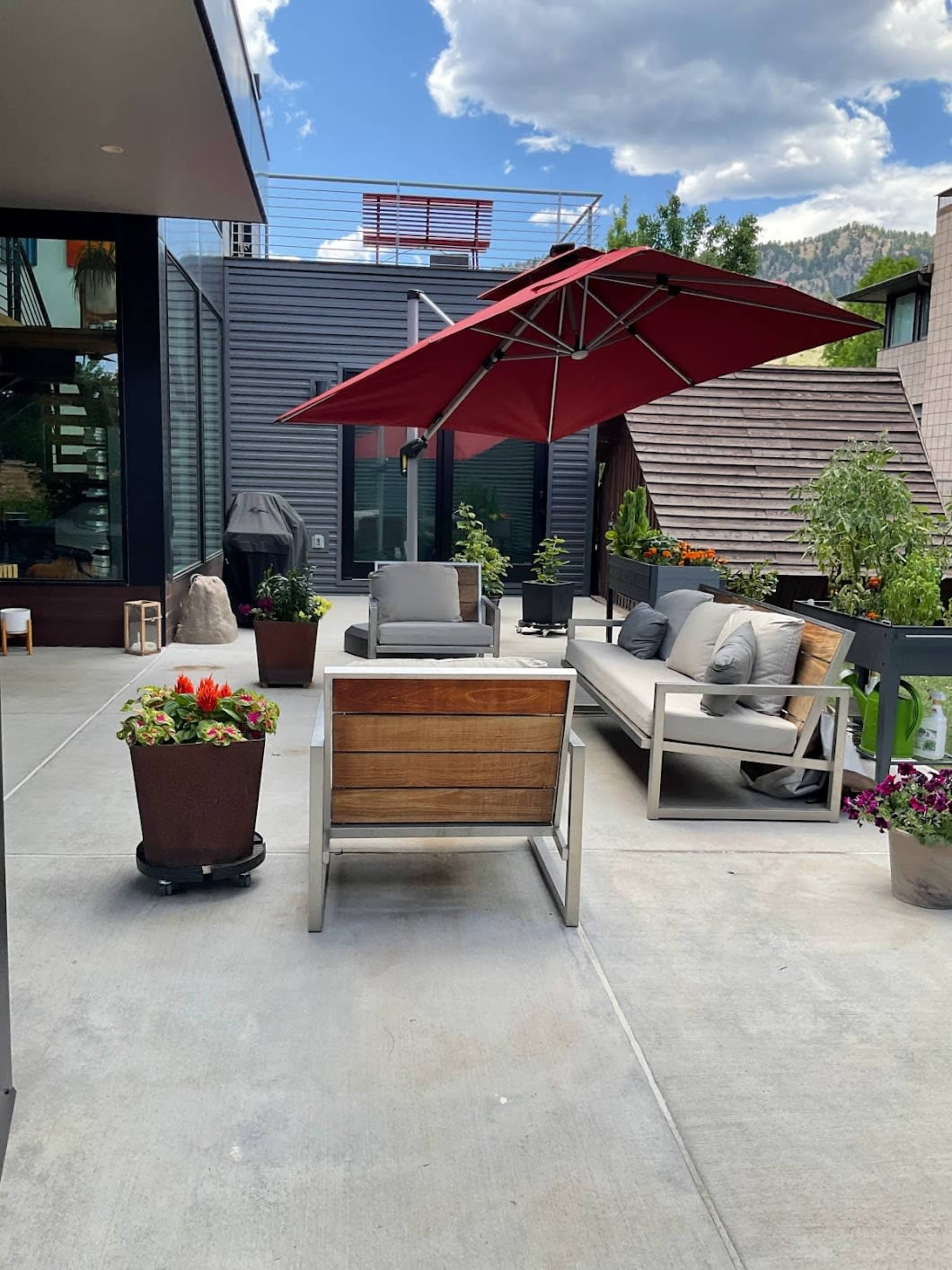 The image shows a modern outdoor patio with a red umbrella, seating areas, and potted plants against a backdrop of buildings and mountains.